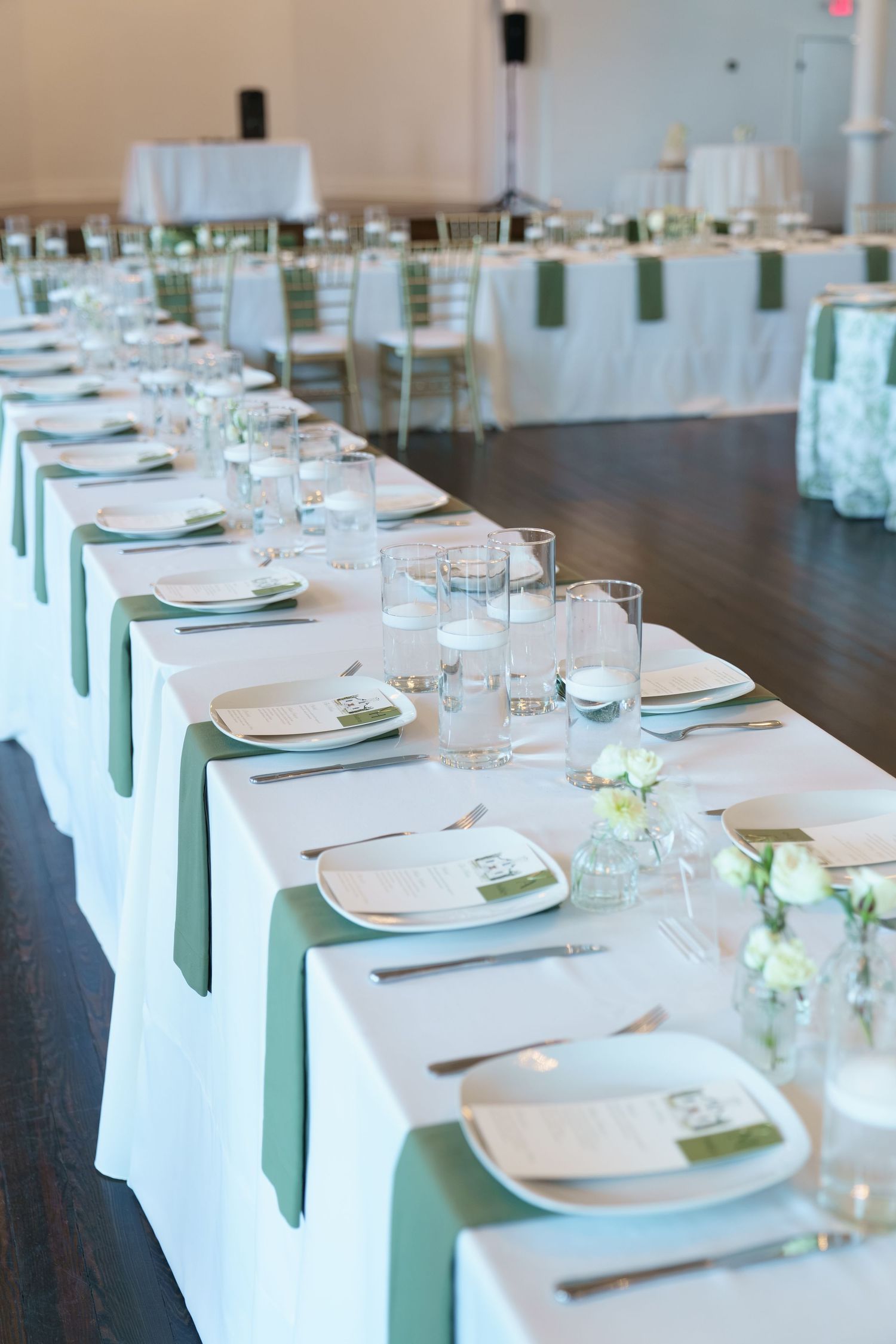 Elegant wedding reception table with white linens, sage green napkins, crystal glassware, and small floral centerpieces.