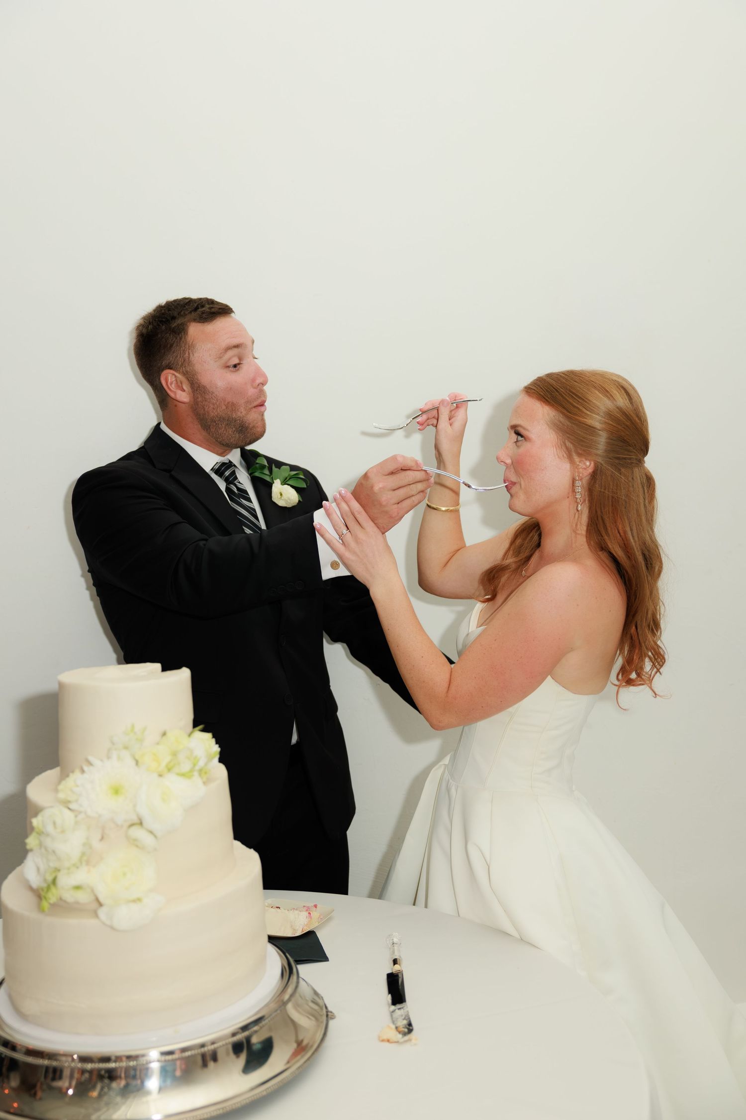 Bride and groom playfully feeding each other wedding cake at their reception beside a white three-tier cake.