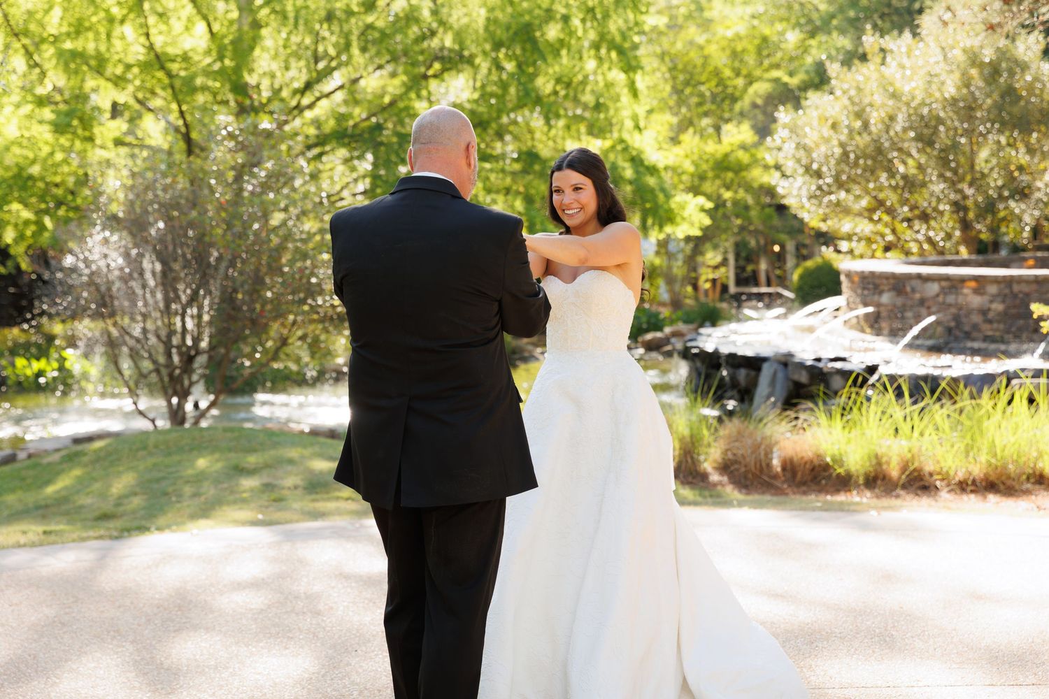 Bride in white strapless gown shares first look moment with groom in black tuxedo outdoors.