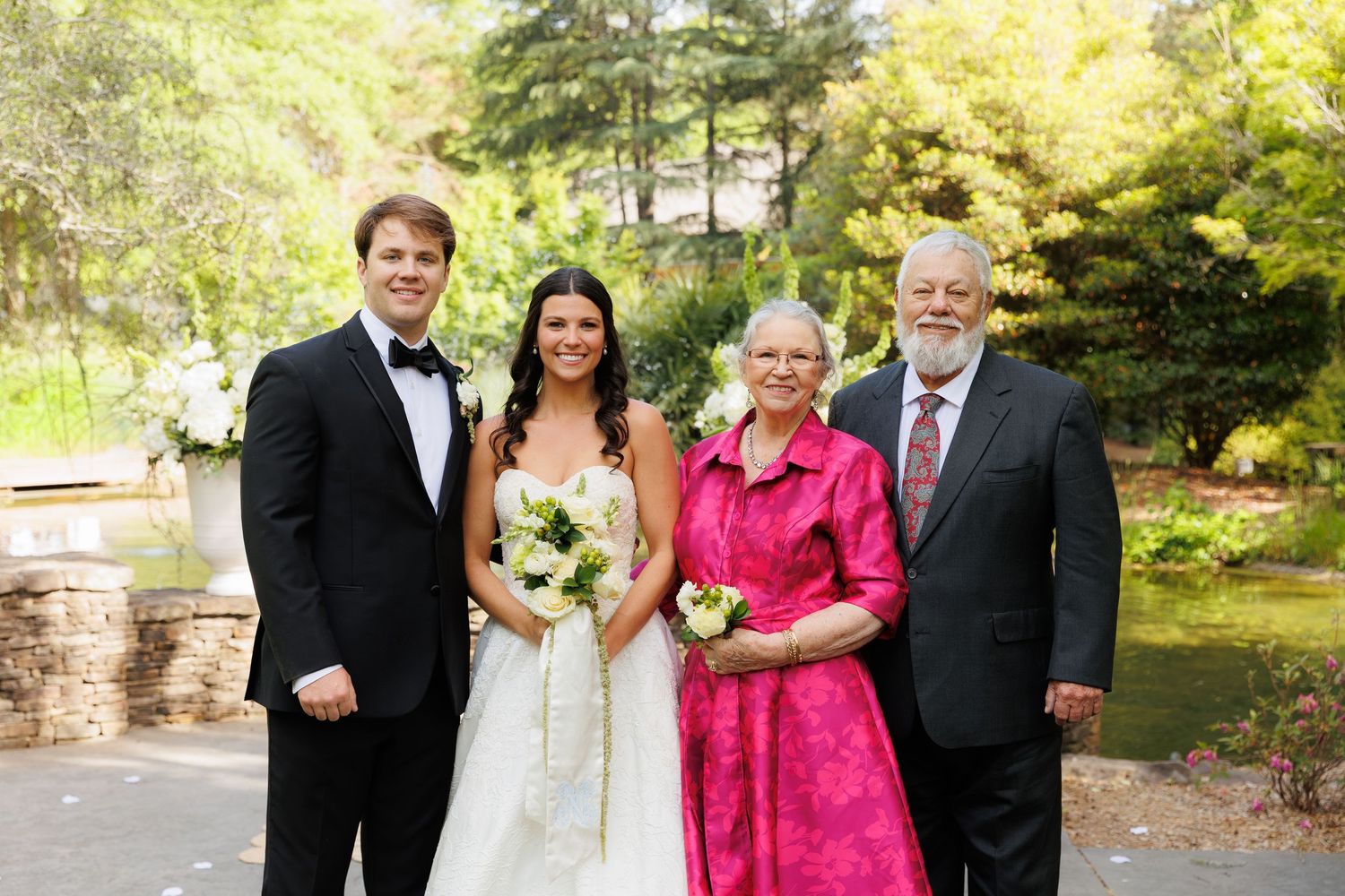 Wedding couple poses with older relatives outdoors, bride in white gown holding bouquet, guests in formal attire.