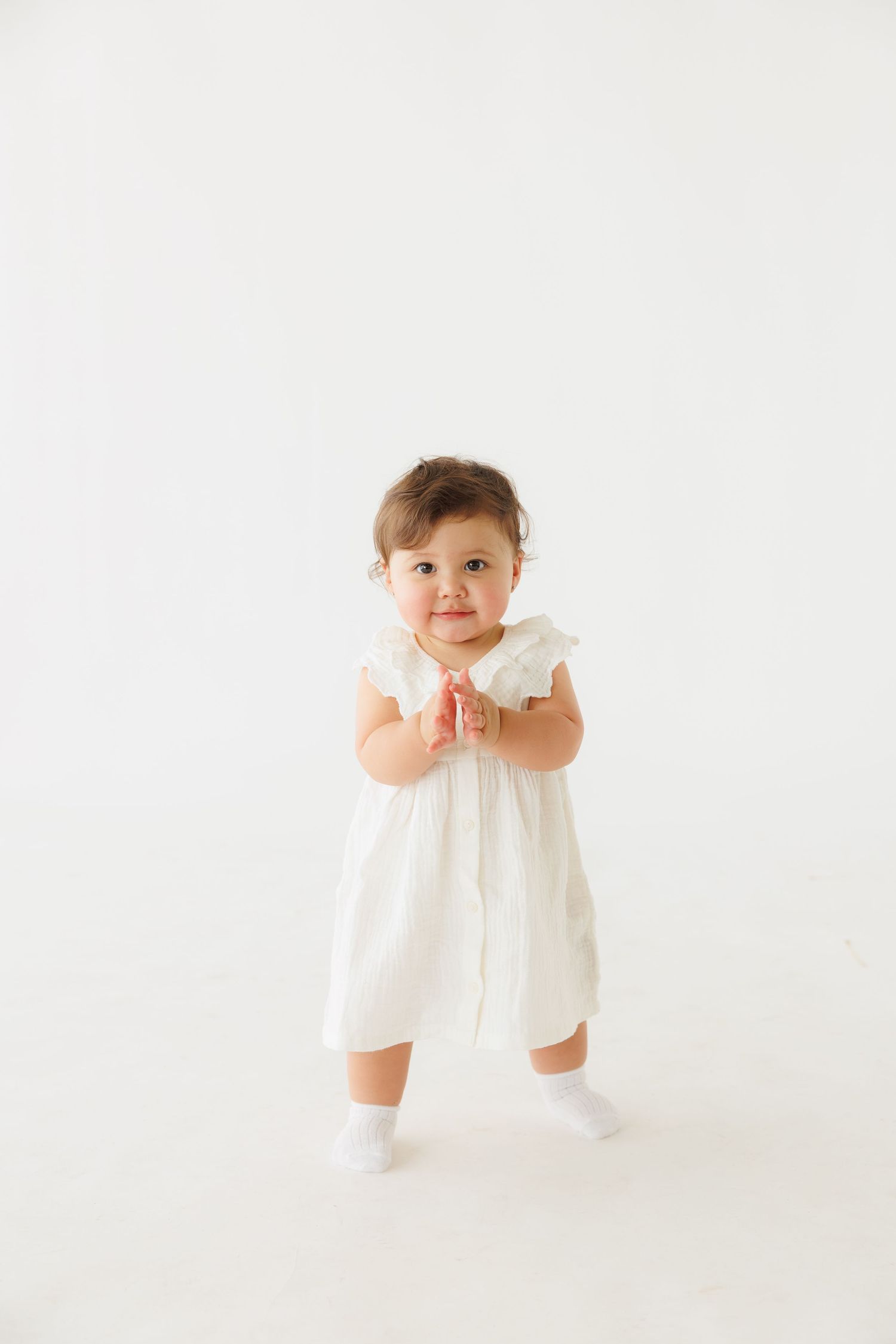 Cute toddler girl in white dress clapping hands on white background.