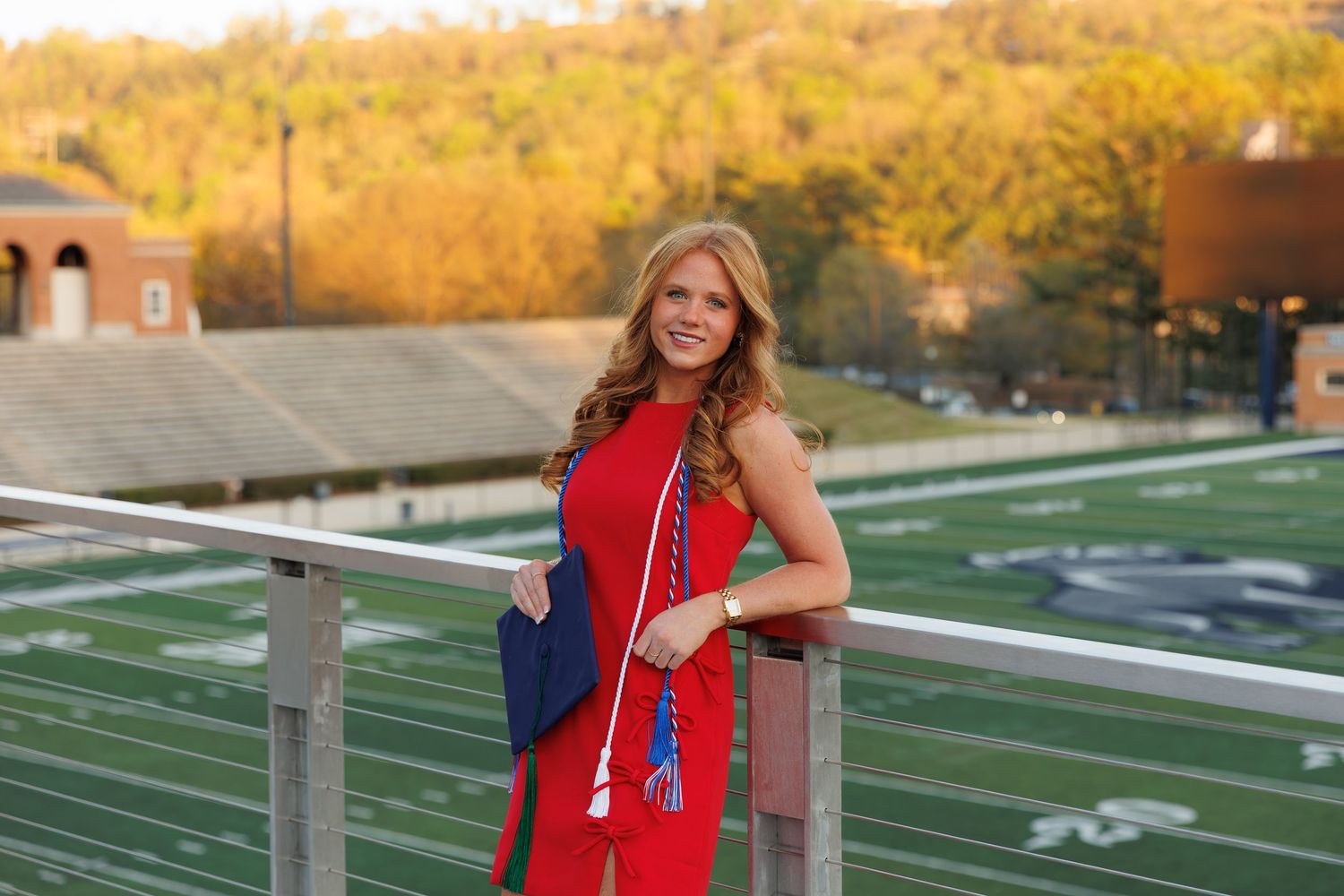 Young woman in red dress holding graduation cap and tassel, posing at college football stadium with green field.
