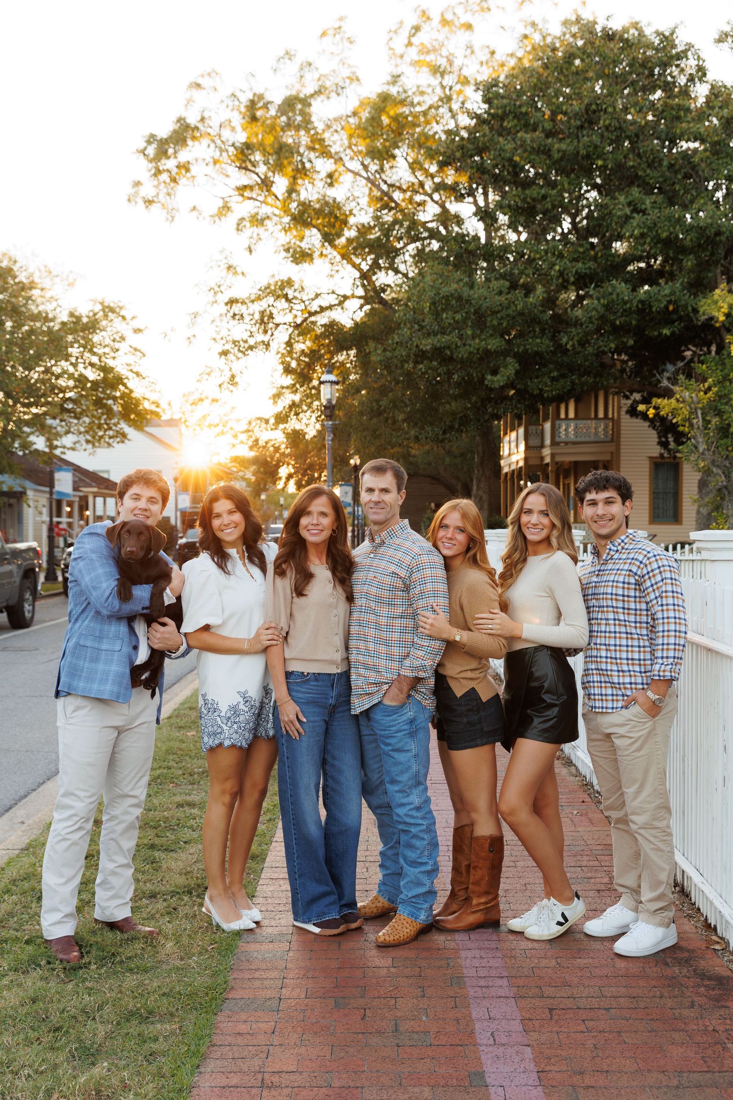 Family of seven poses with a dog on a brick sidewalk at golden hour, dressed in casual fall outfits.