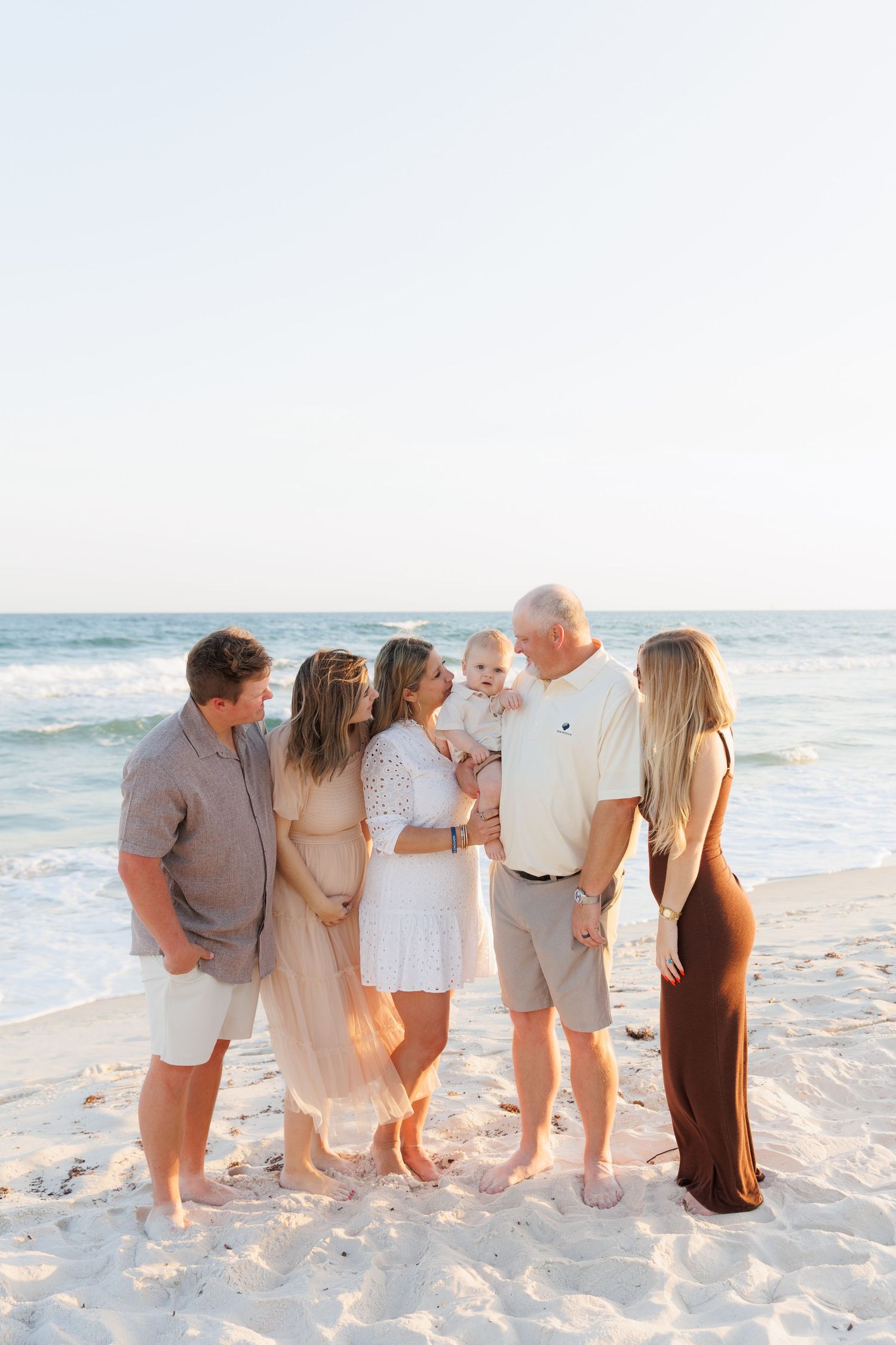 Family of six poses on sandy beach with ocean waves, wearing coordinated neutral outfits during golden hour.
