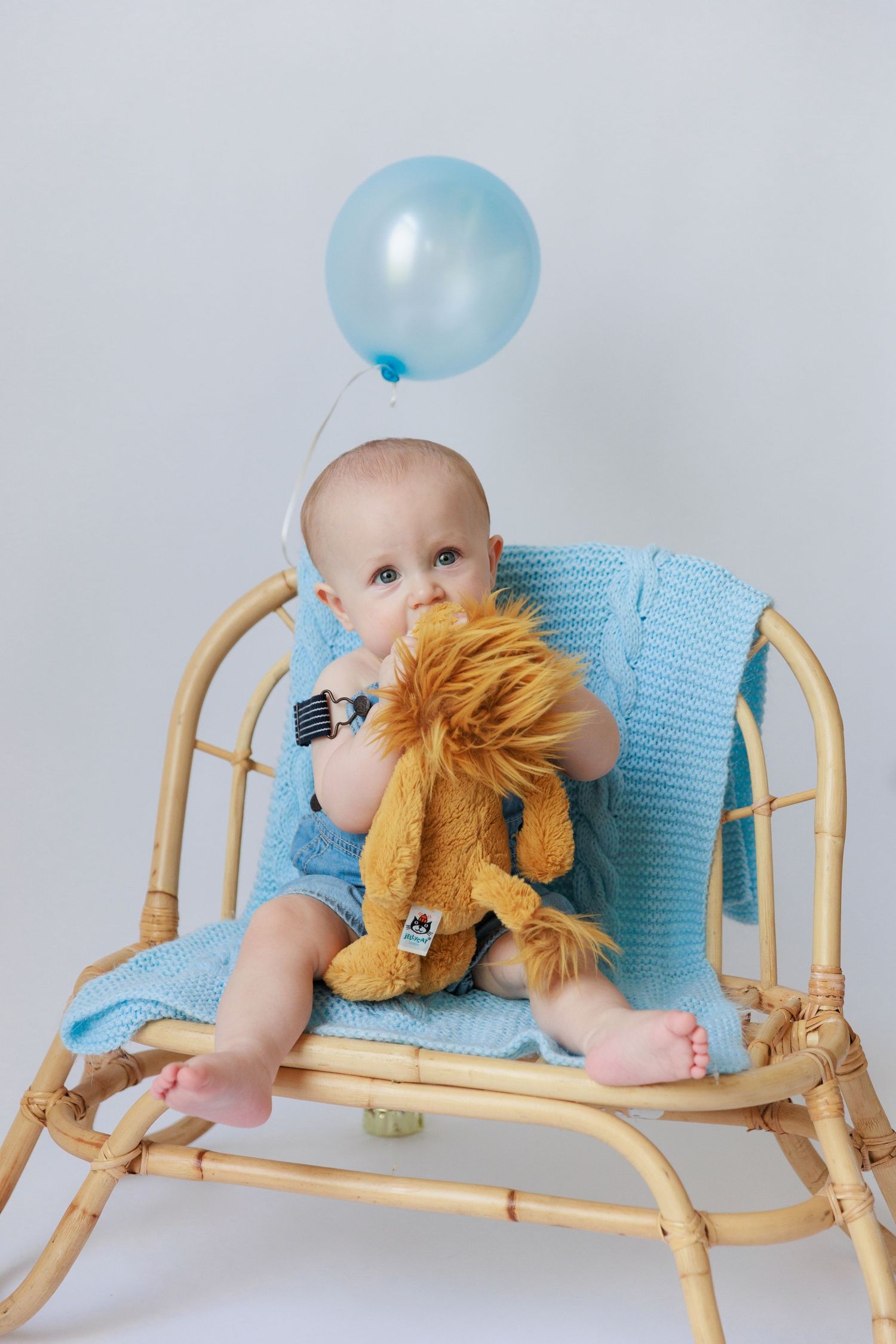 Baby in light blue outfit plays with plush lion toy on bamboo lounger while blue balloon floats nearby.