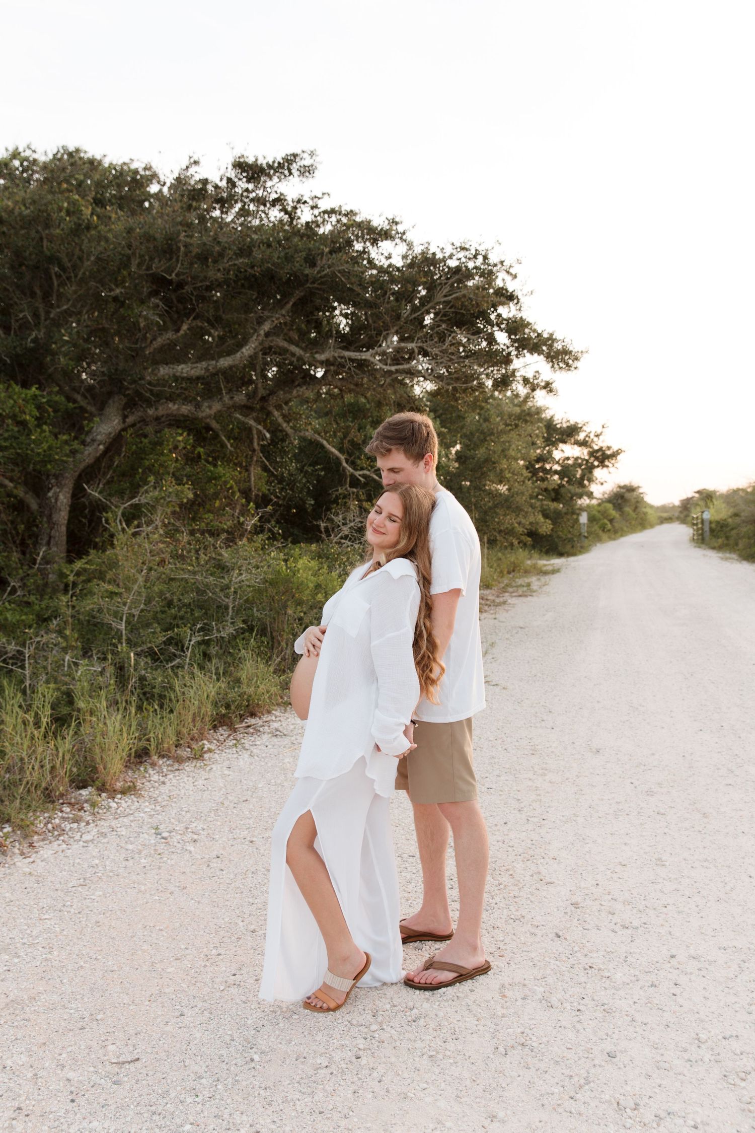 A couple embraces on a sandy beach path at sunset with trees and greenery in the background.