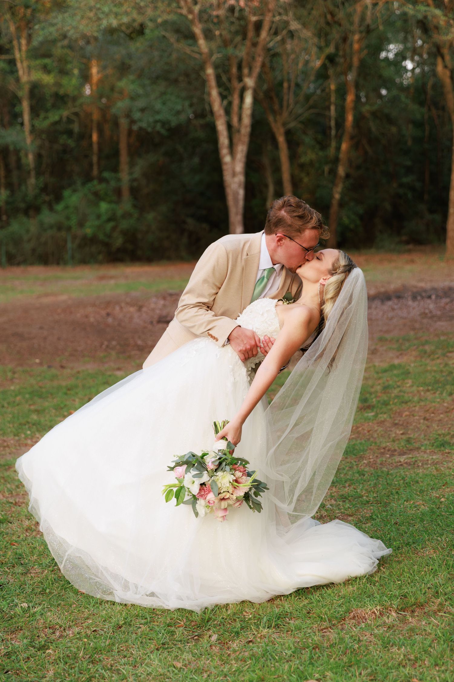 A couple in wedding attire shares a romantic dip pose on a grassy lawn surrounded by trees.