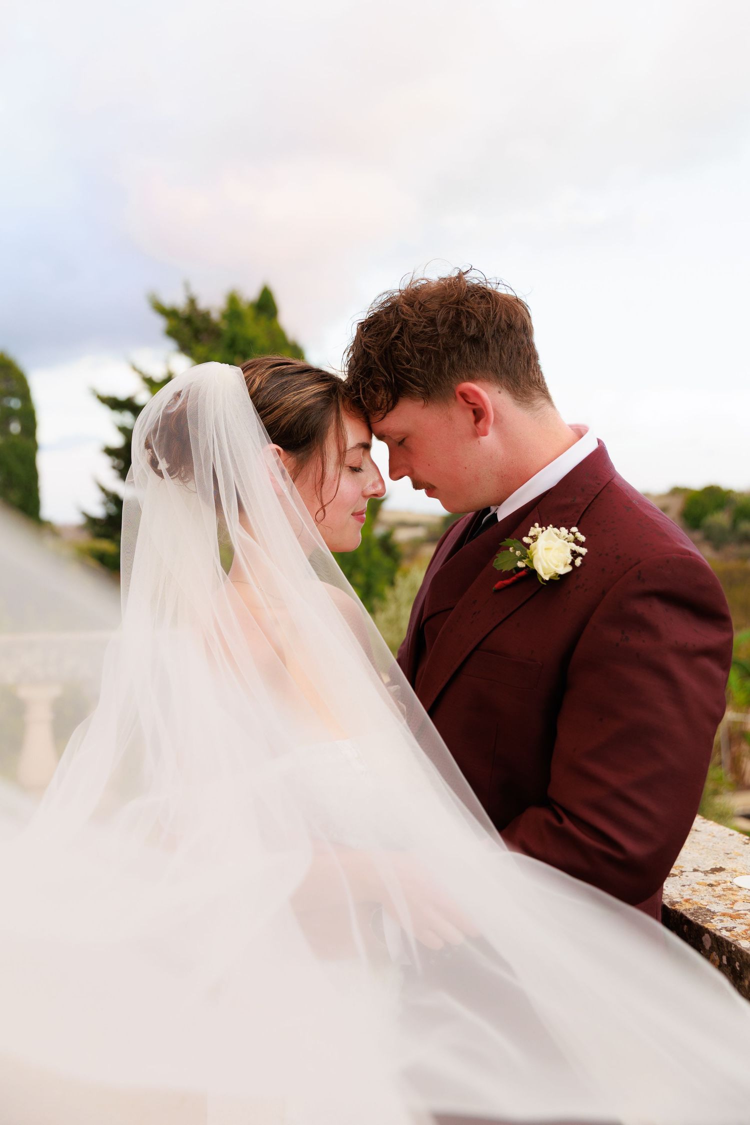 A romantic wedding moment with flowing veil and burgundy suit against a soft sky.
