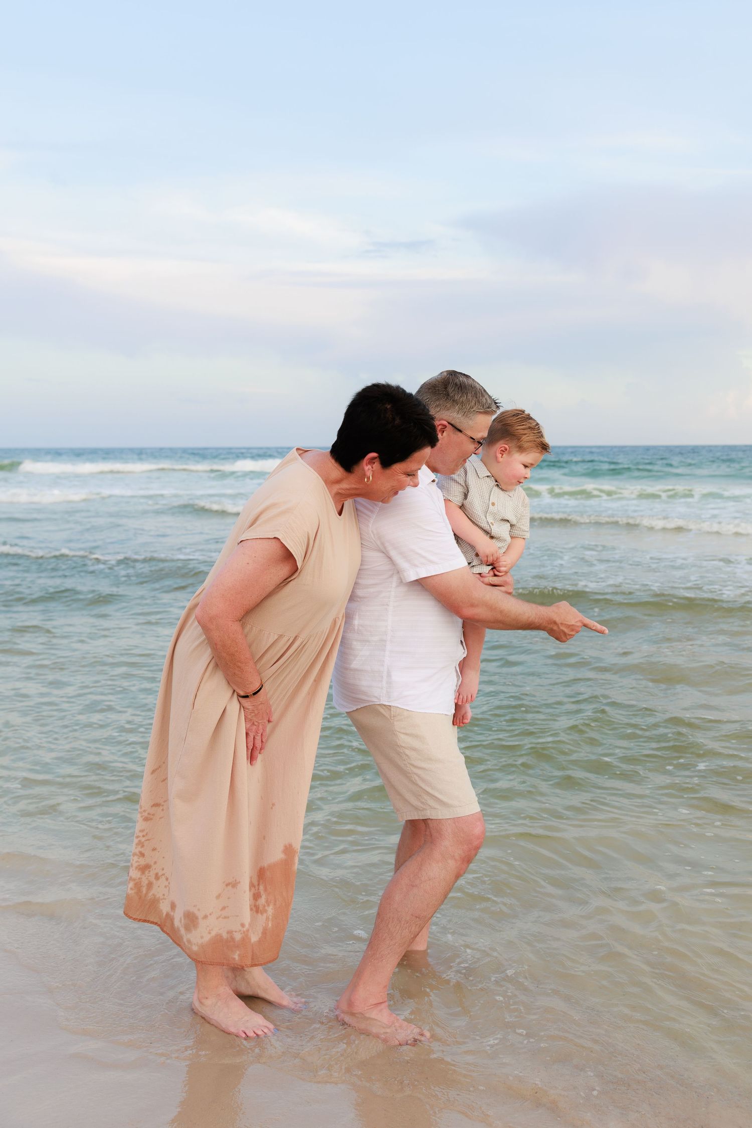 Three people standing at the edge of the ocean waves on a beach, looking down at the water together.