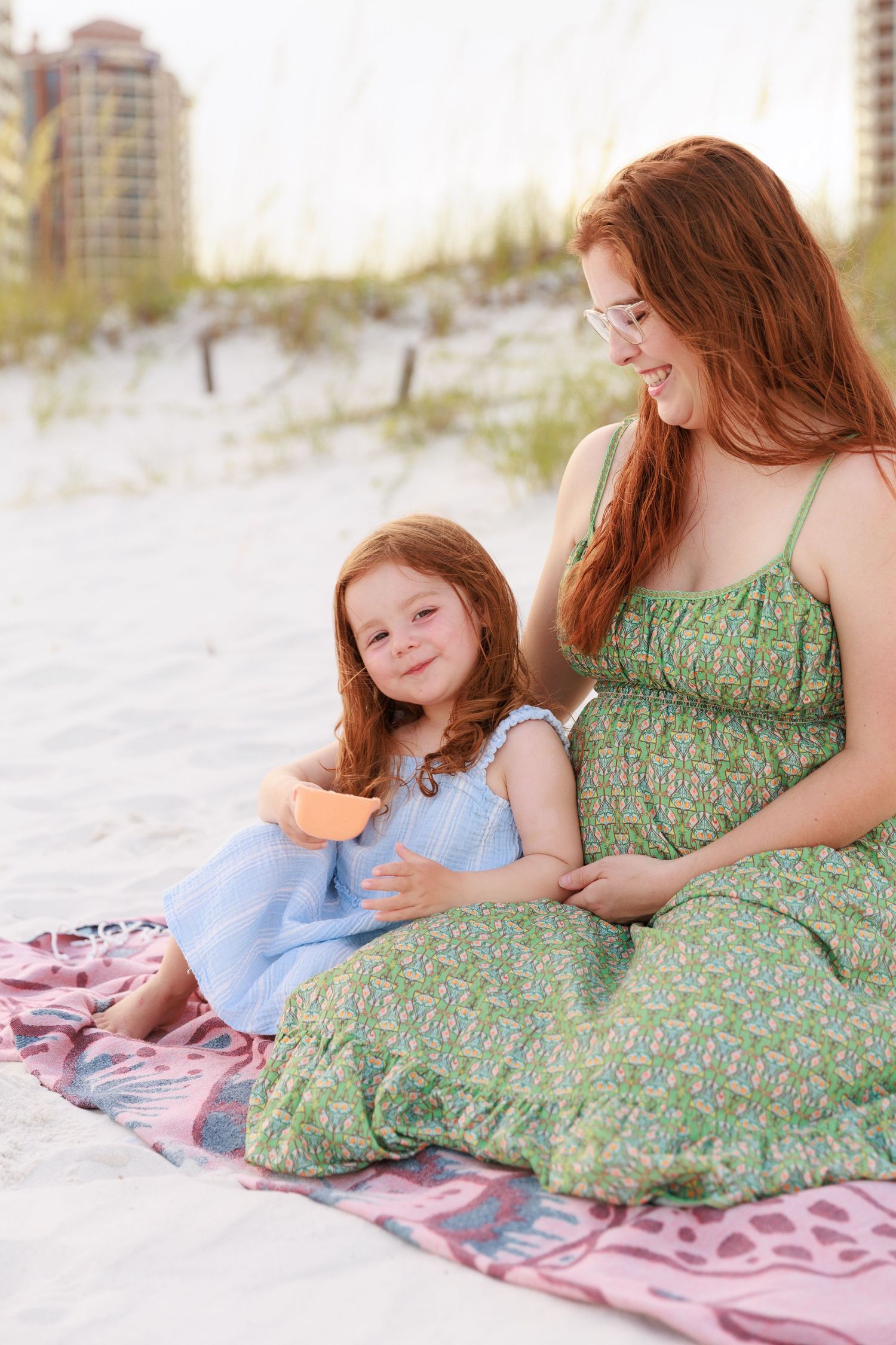 A parent and child share a tender moment while relaxing together on a pink blanket at the beach.