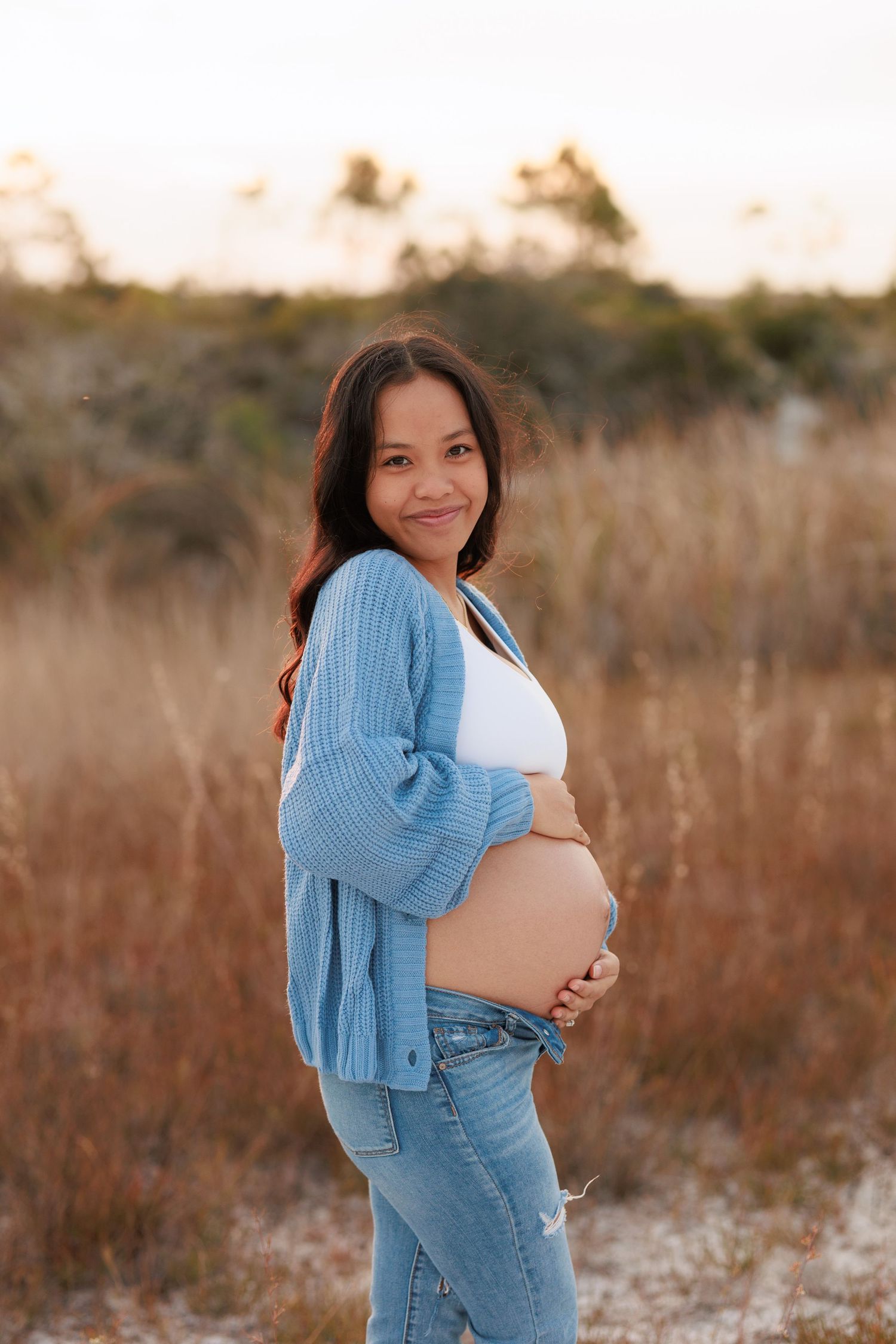 Expectant mother in blue cardigan and jeans poses outdoors during golden hour maternity session.