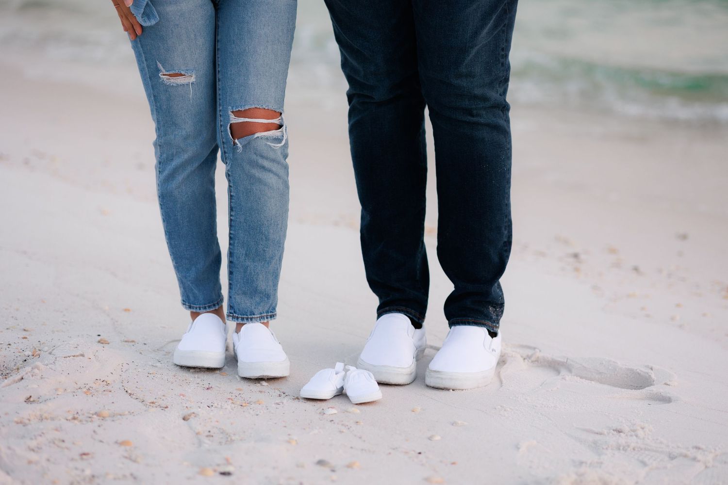 Close-up view of legs wearing distressed blue jeans and white sneakers walking on a sandy surface.