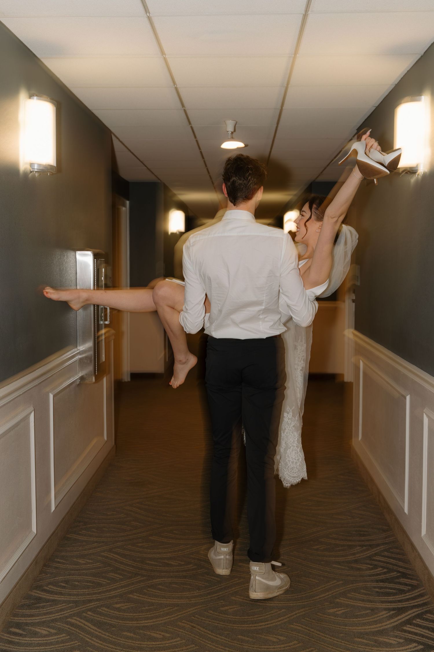 A couple in white wedding attire dances playfully in a modern hotel hallway with carpeted floors.