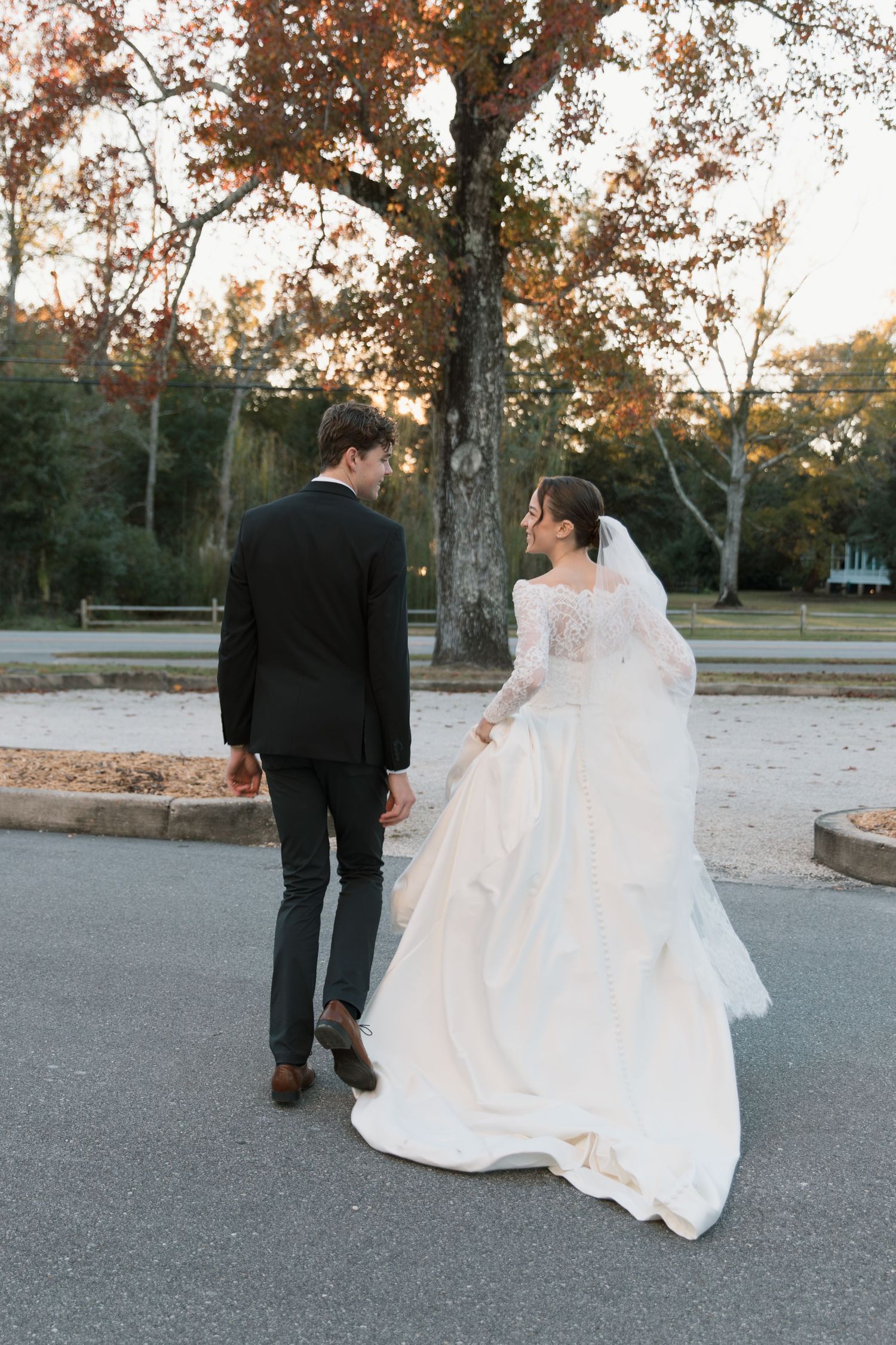 Wedding couple walking together on a paved path surrounded by autumn trees.