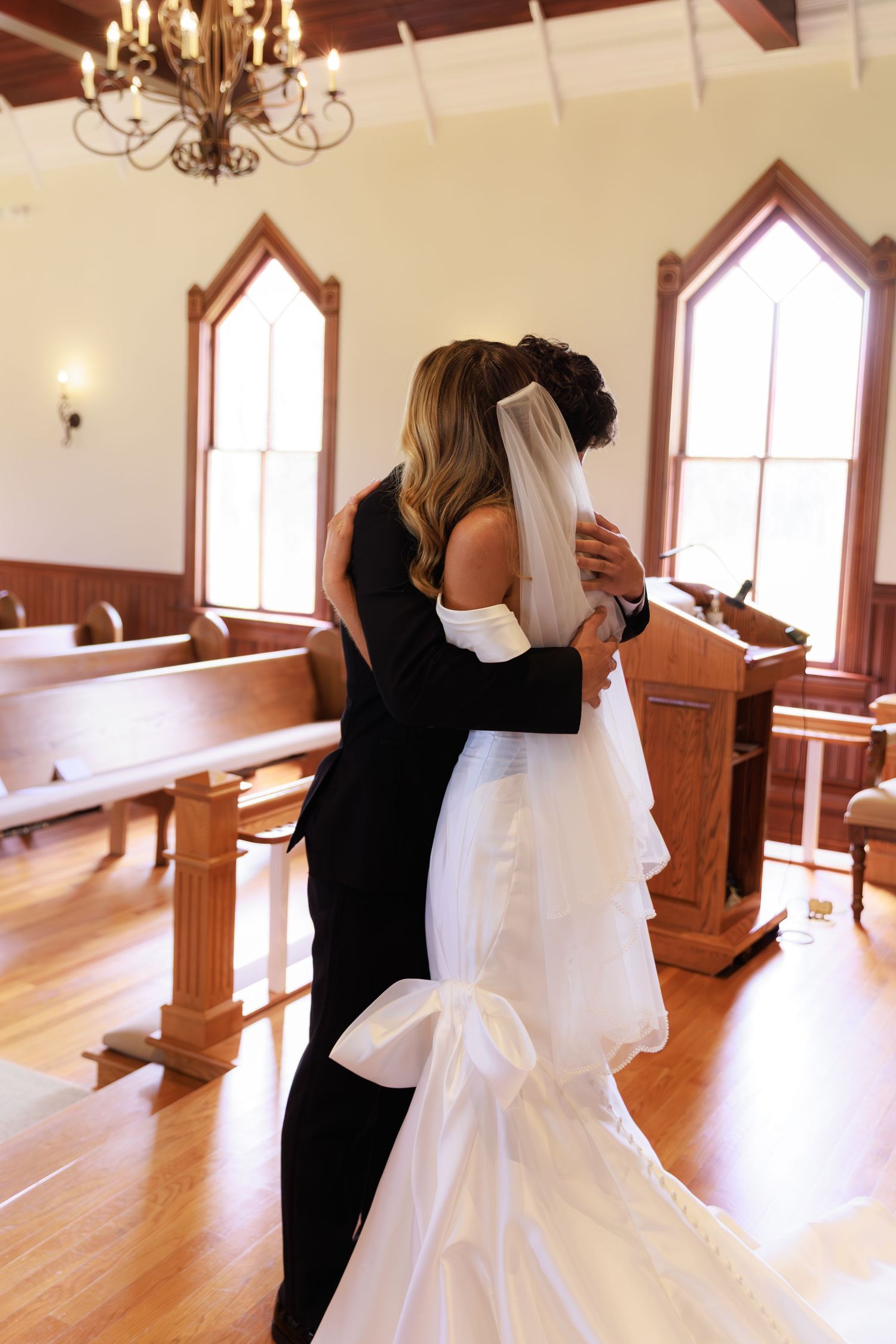 An emotional embrace between two people in a church sanctuary with wooden pews and gothic arched windows.