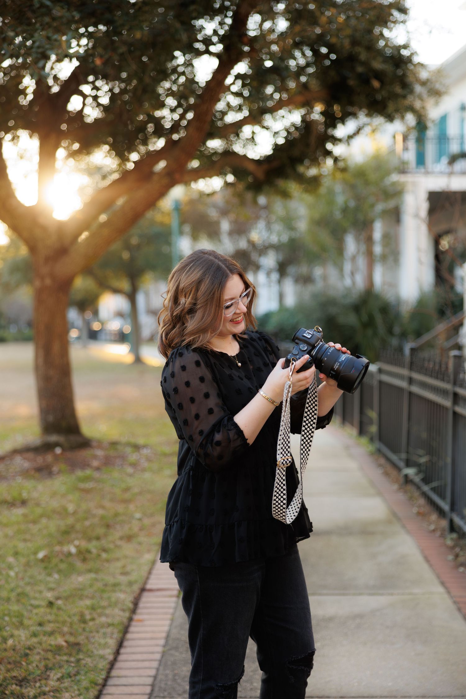 Woman in black outfit smiling while reviewing photos on DSLR camera outdoors at golden hour.