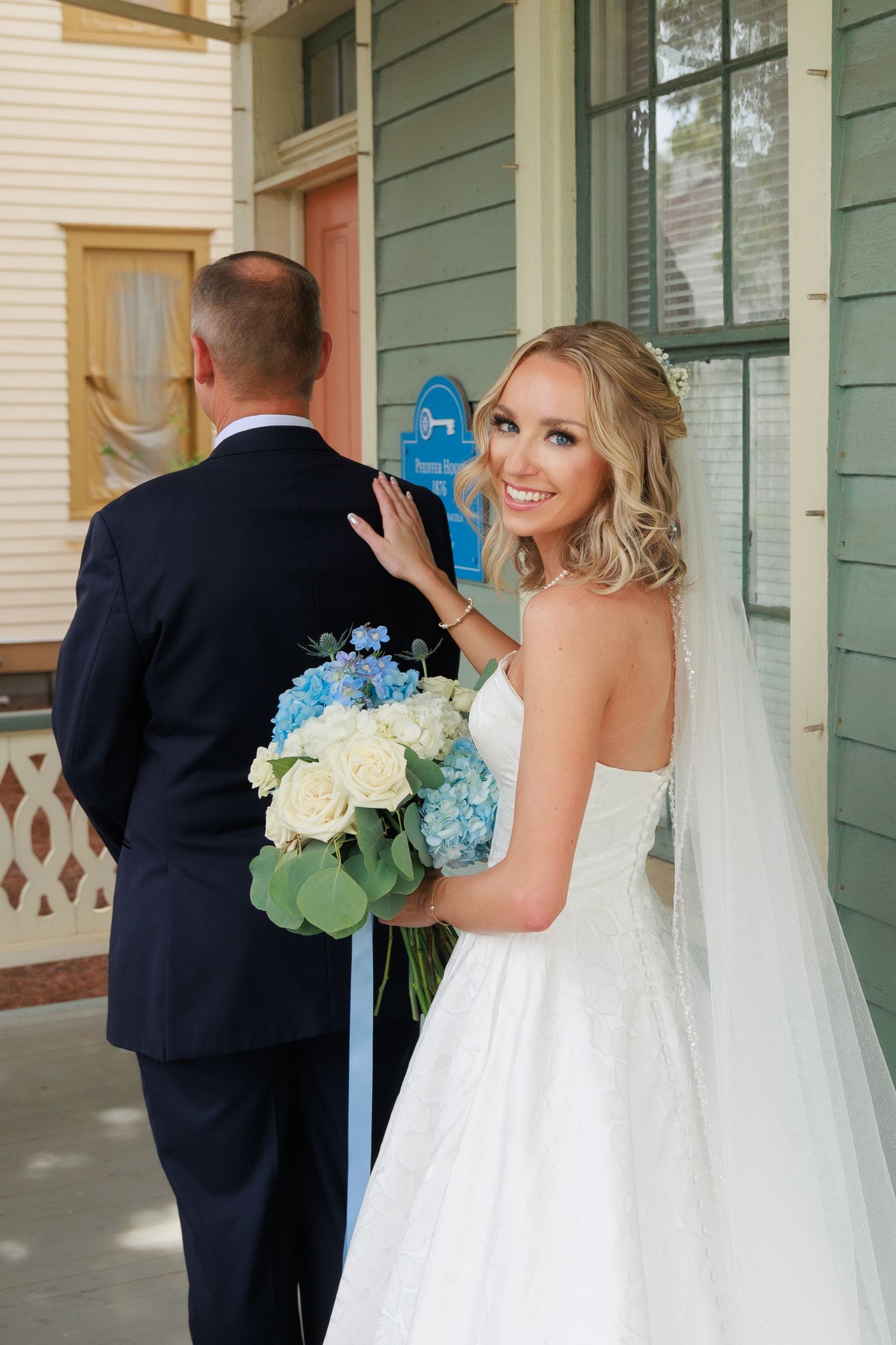 Bride in white dress holds bouquet while groom faces away during first look photo.