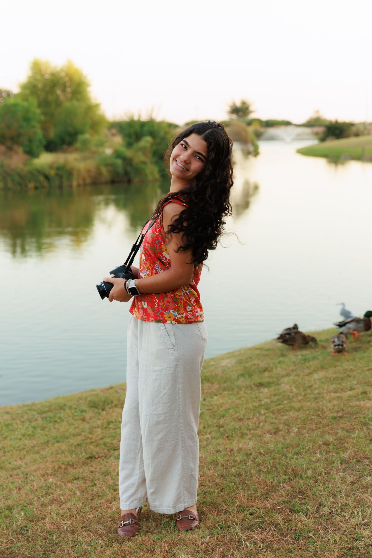 Photographer in bright summer attire poses with camera by waterfront with fountain view.