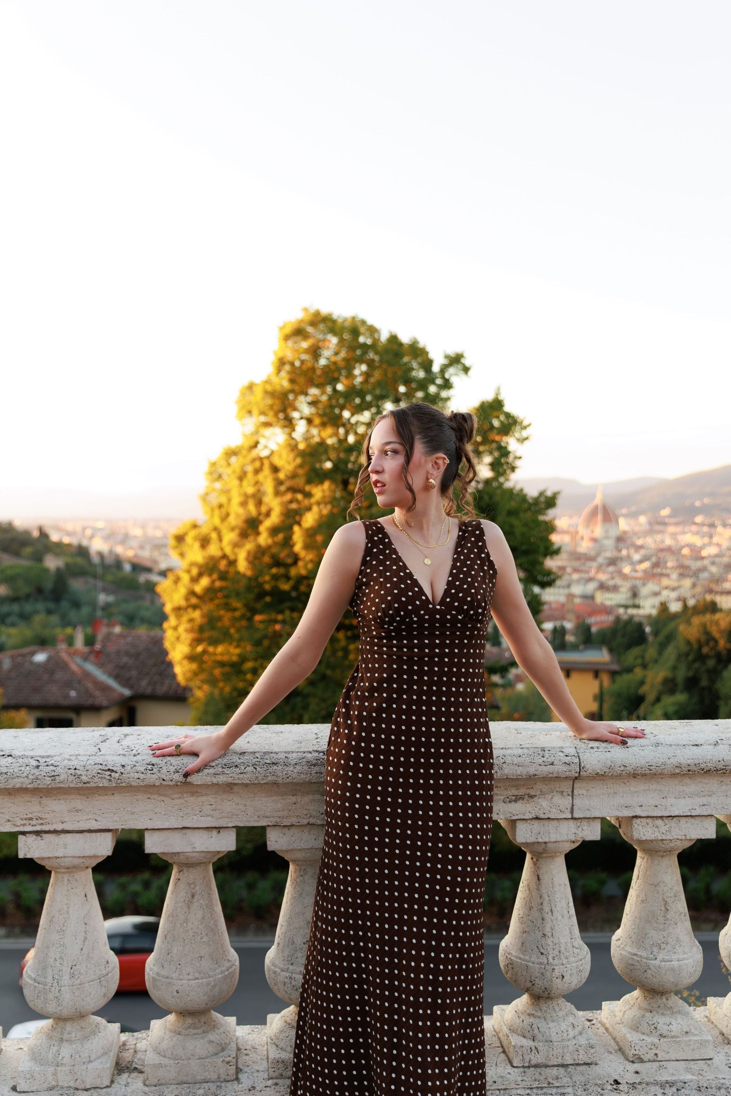 Young woman in polka dot maxi dress poses at stone balustrade with Florence cityscape at sunset.