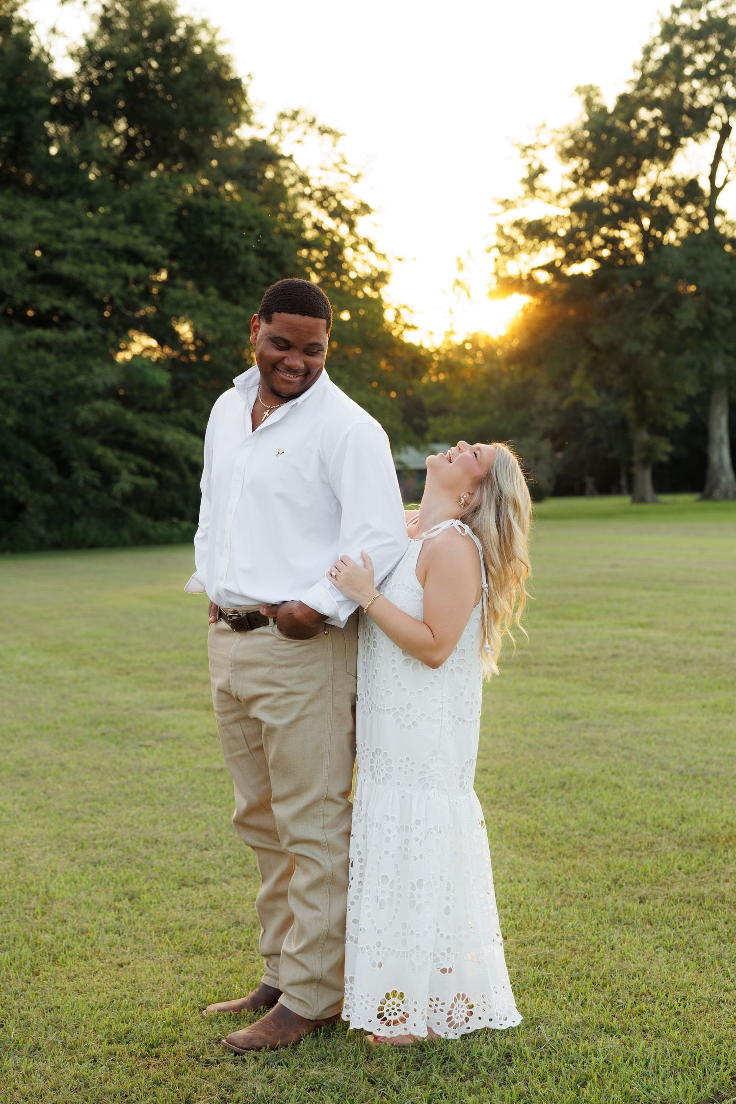 Couple laughing together at golden hour sunset in a green park, man in white shirt and woman in white lace dress.
