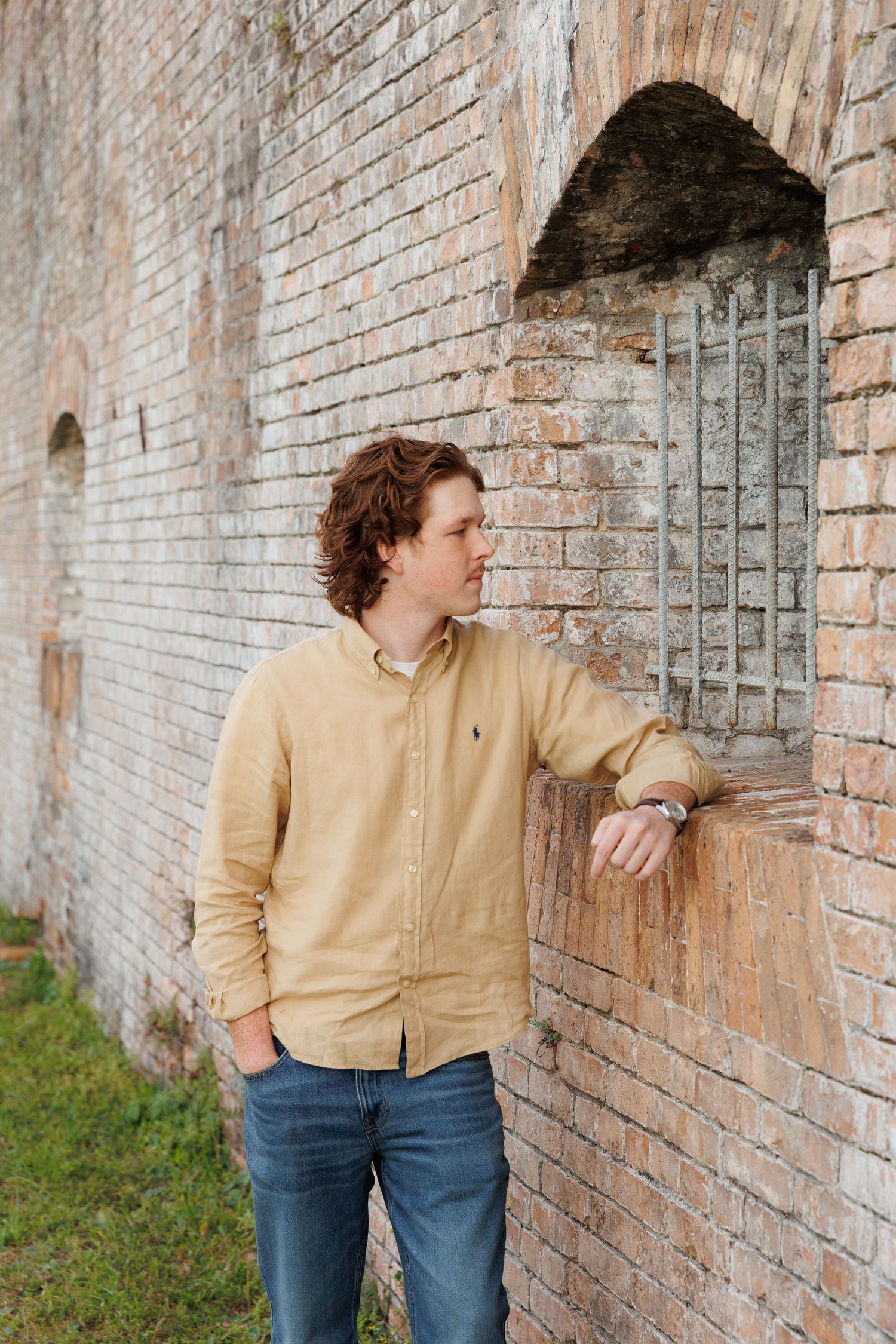 Young man in yellow Ralph Lauren shirt leans against rustic brick wall with barred window, wearing jeans and a watch.