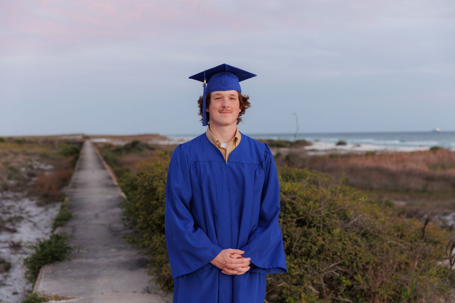 A smiling graduate in blue cap and gown stands on a beach boardwalk at sunset for senior portrait photos.