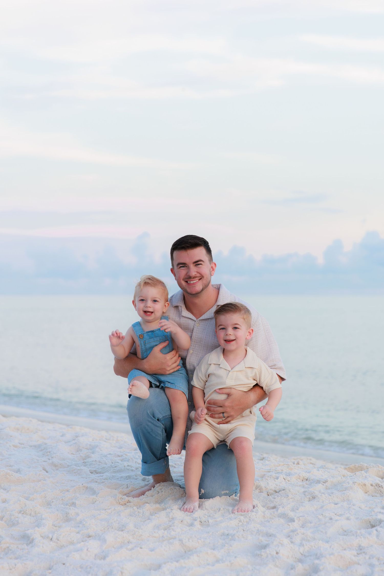 A smiling father kneels on a white sand beach holding two young boys with calm ocean waters behind them.