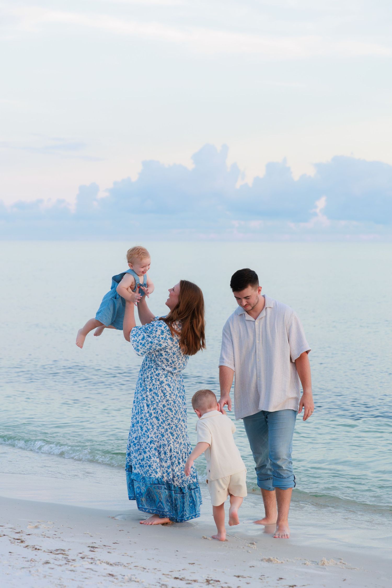 Family of four enjoying a beach walk at sunset, mother lifting baby girl while toddler walks with father.