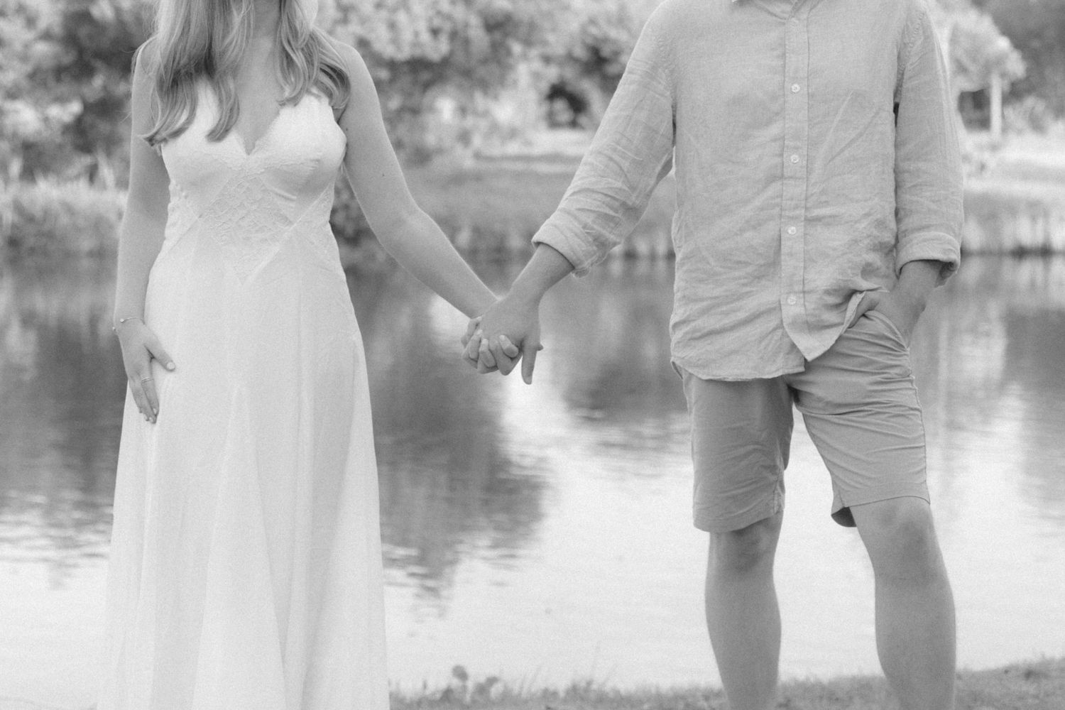 Black and white photo of a couple holding hands by a lake, woman in white dress, man in casual shirt and shorts.