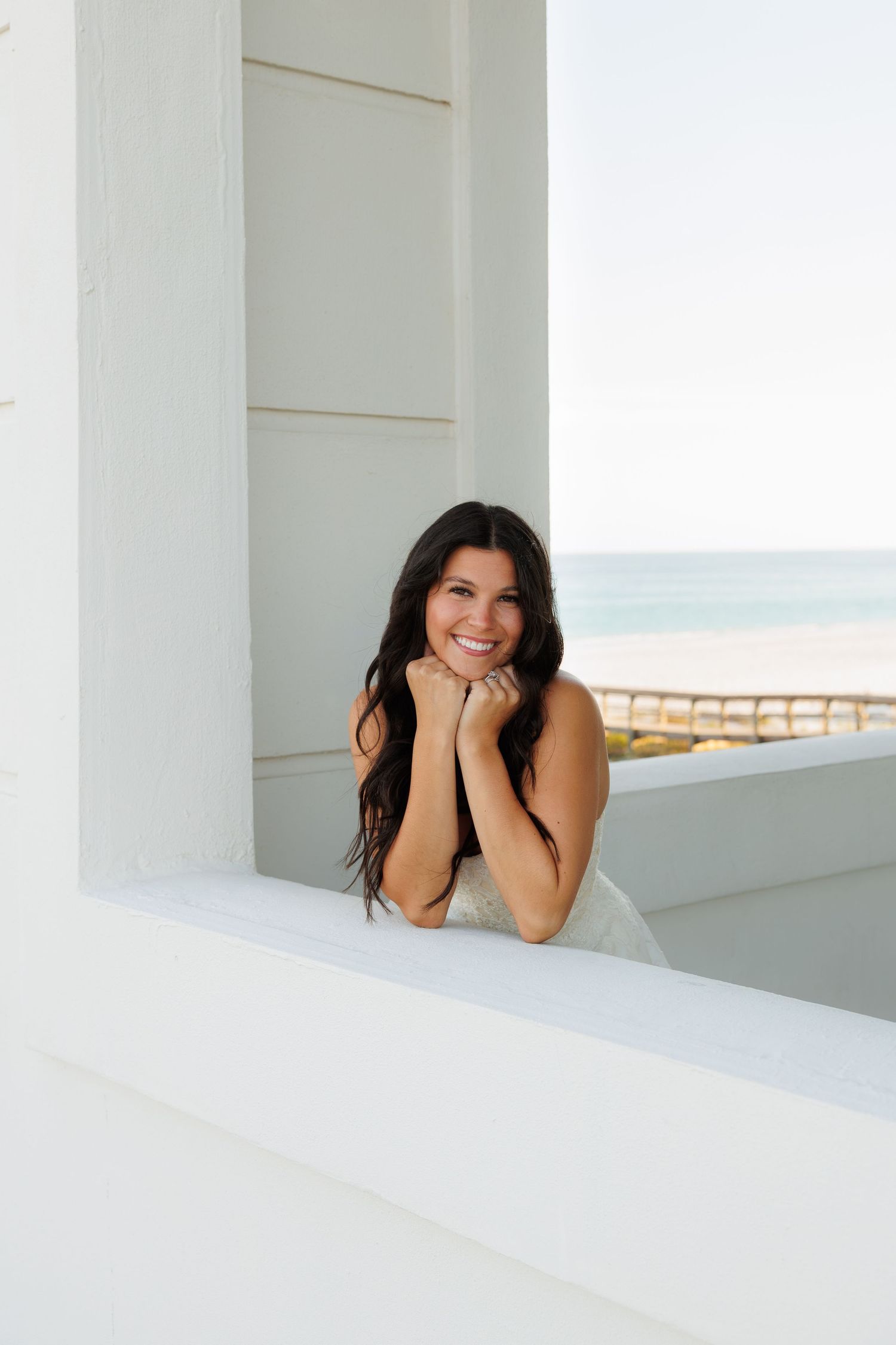 Smiling bride with dark hair leans on white ledge with ocean view behind her.