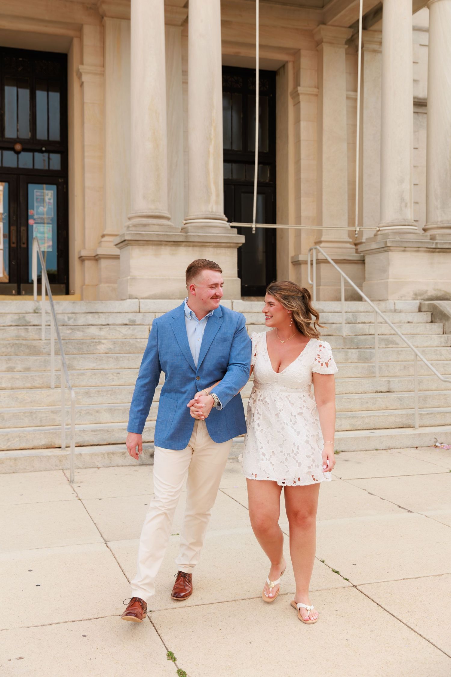 Happy couple holding hands and walking in front of a grand stone building, smiling at each other.