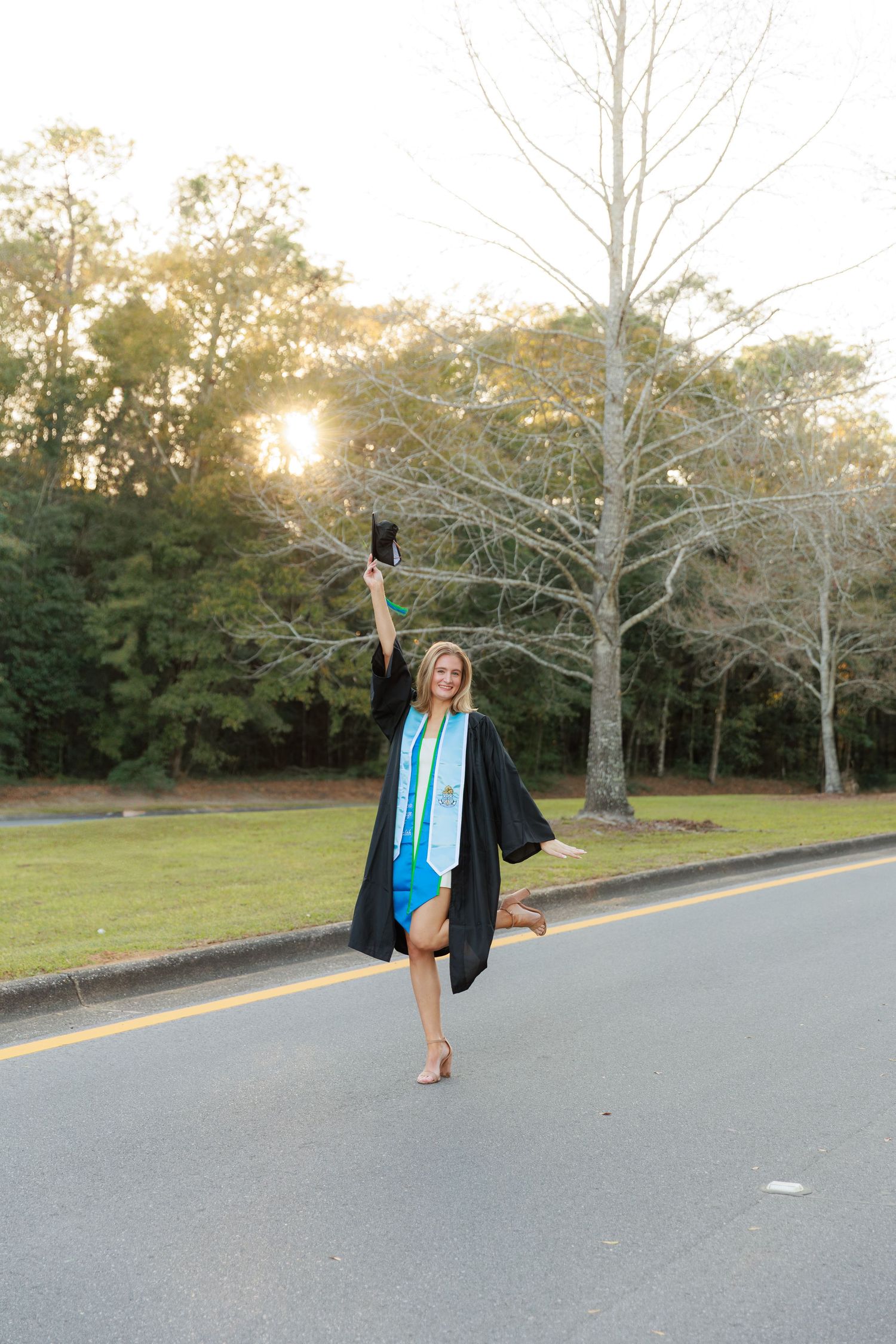 A graduate in cap and gown poses joyfully on an empty road during a sunset photoshoot session.