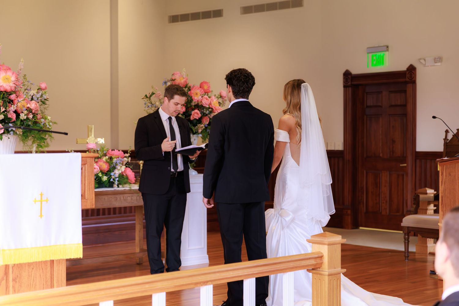 A wedding ceremony takes place in a church sanctuary with pink floral arrangements and wooden pews.