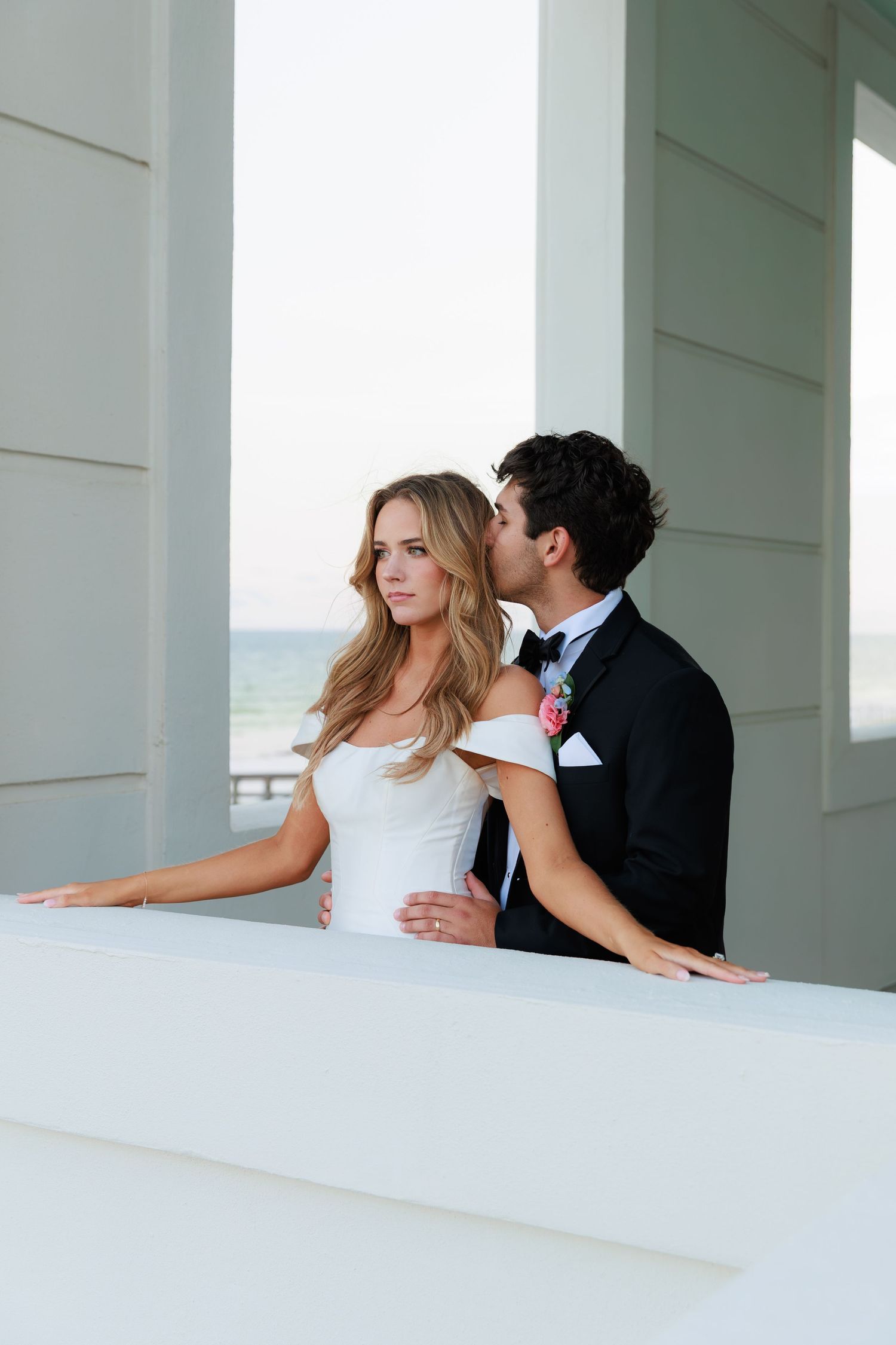 Romantic wedding couple embracing on white balcony overlooking ocean view.