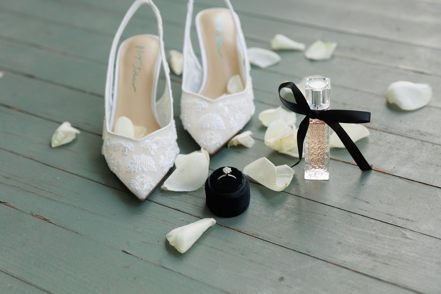 White lace bridal shoes and scattered rose petals arranged on a rustic wooden floor.