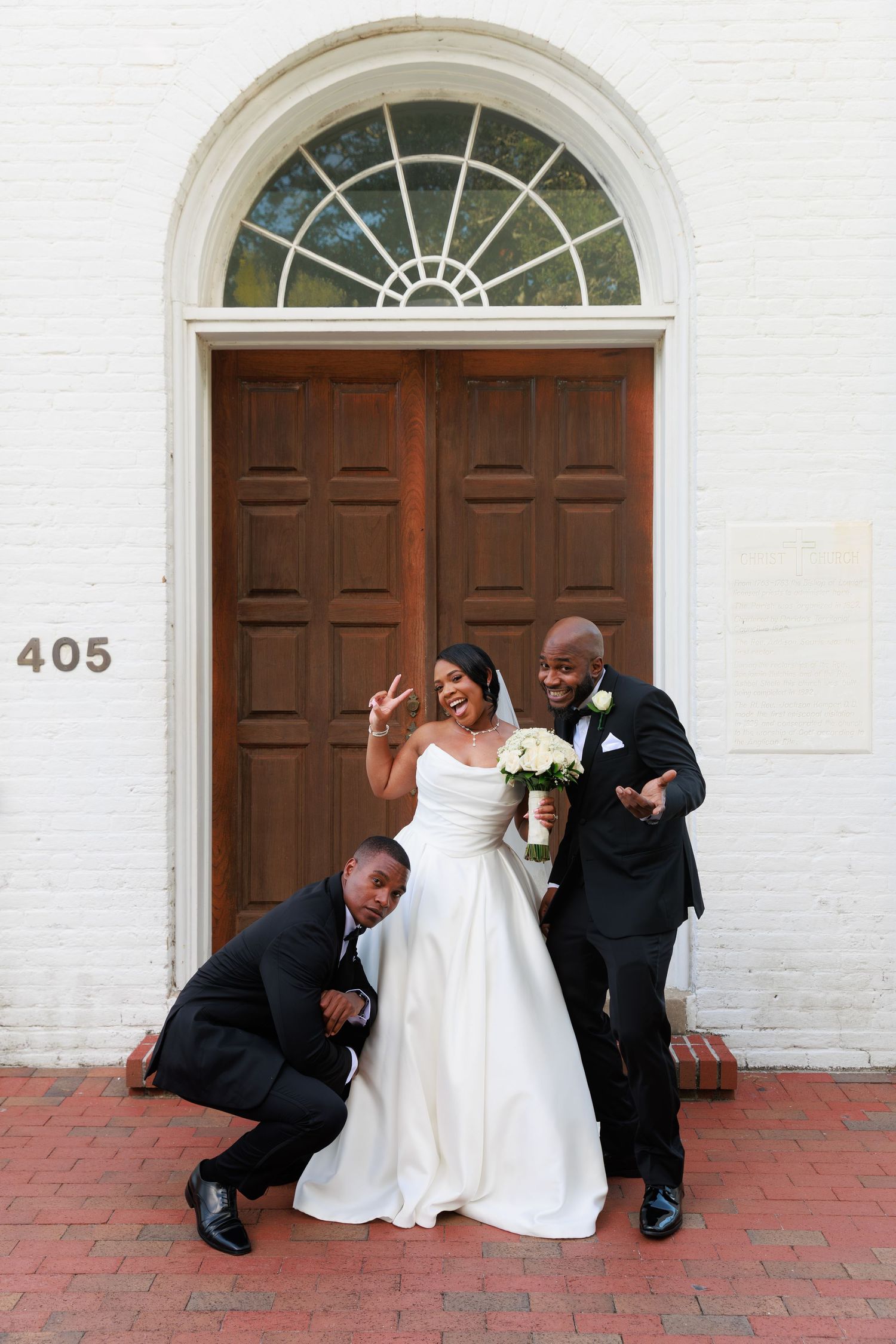 Newlyweds and wedding party pose playfully in front of a historic white church with brown wooden doors.