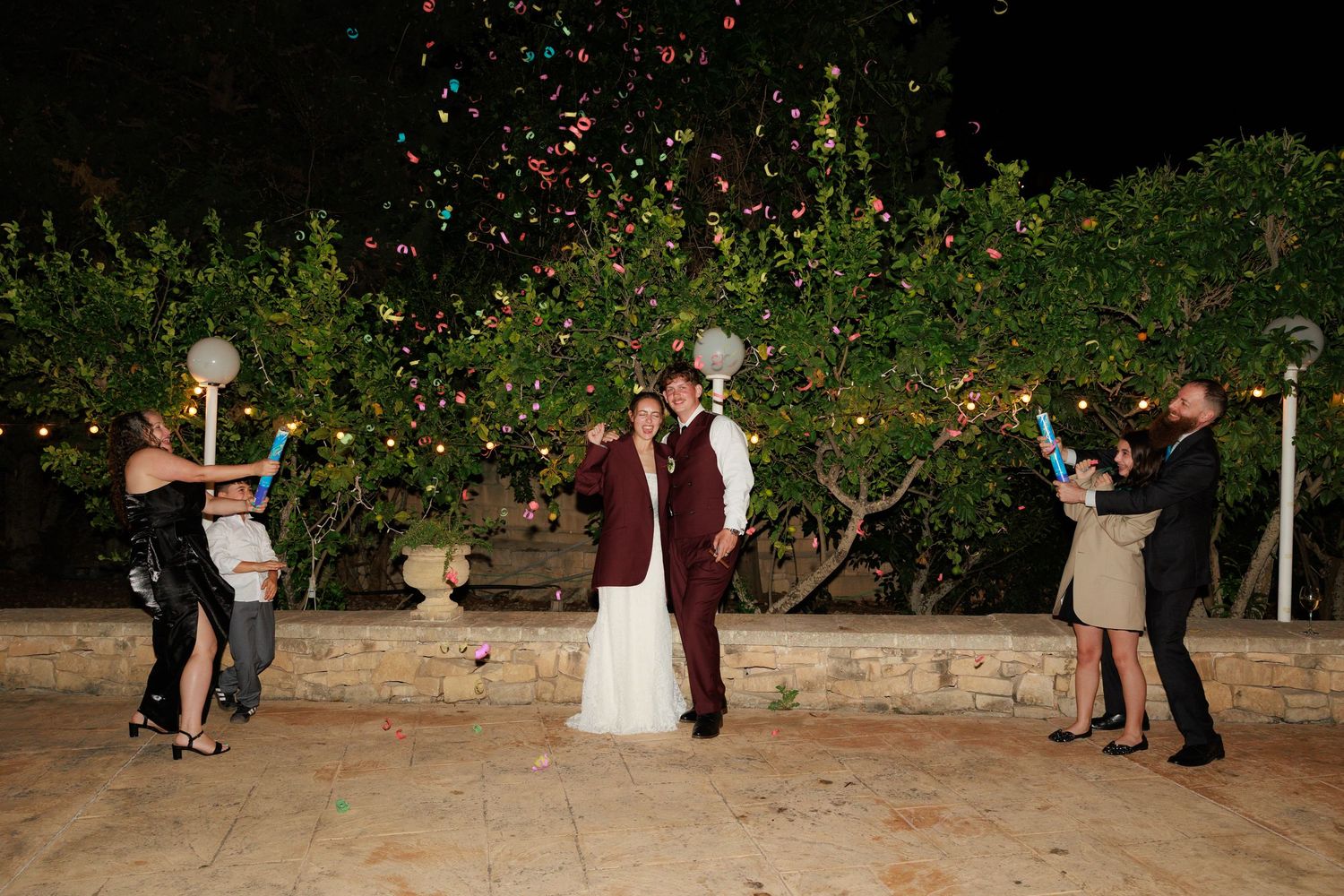 Wedding couple poses while guests throw flower petals in garden setting at night.