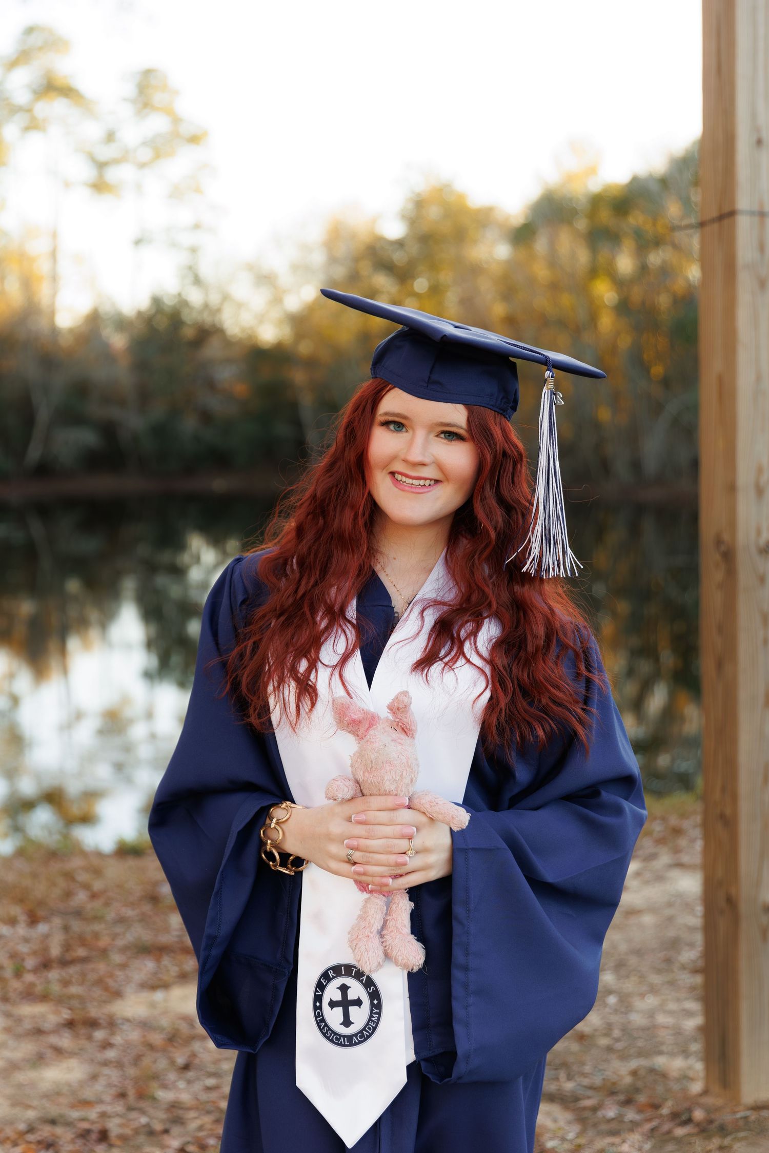 A graduate in navy blue cap and gown poses outdoors near a lake during a graduation photoshoot.