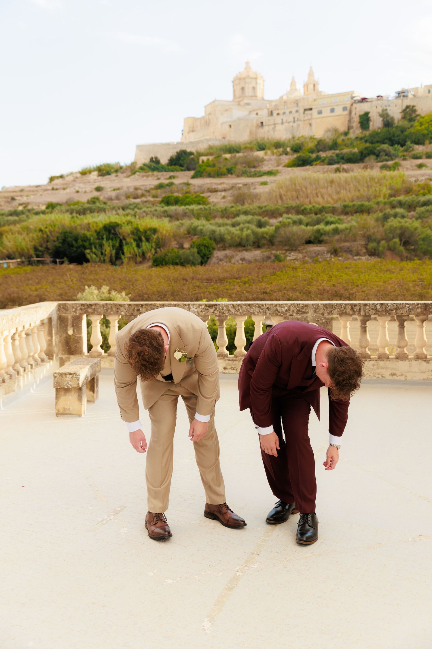 Two people in formal attire bend down to examine something on the ground with a Mediterranean castle in the background.