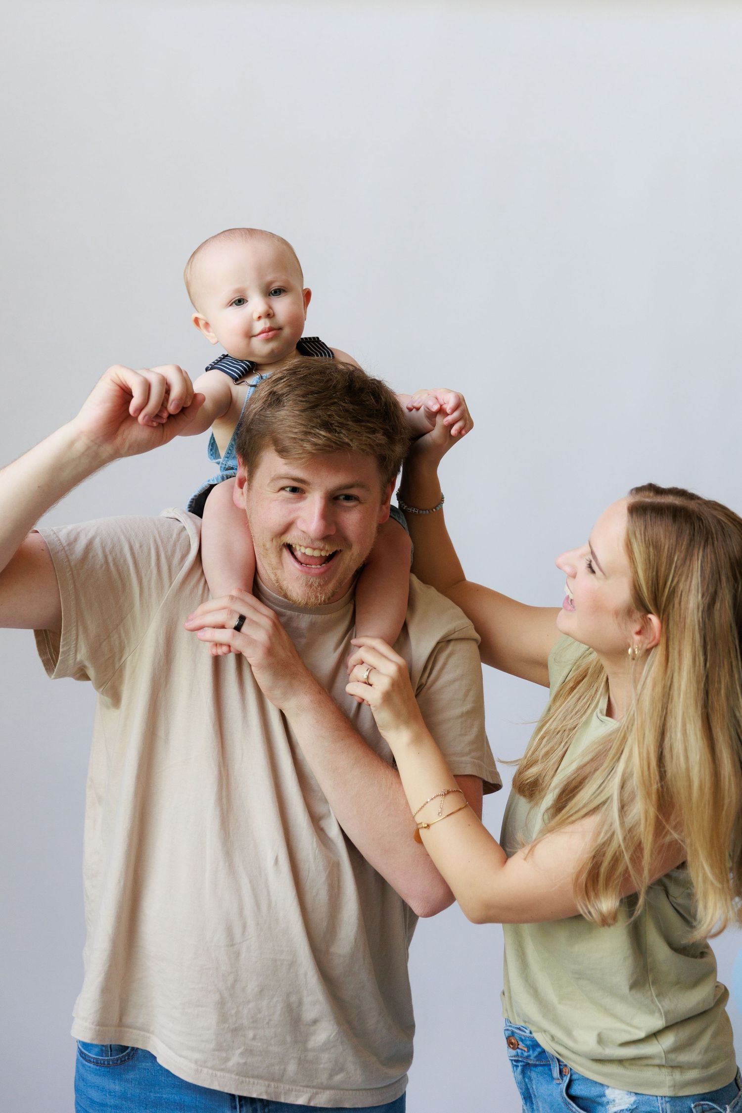 A joyful family moment captured as a baby rides on shoulders while a couple shares laughter against a light background.