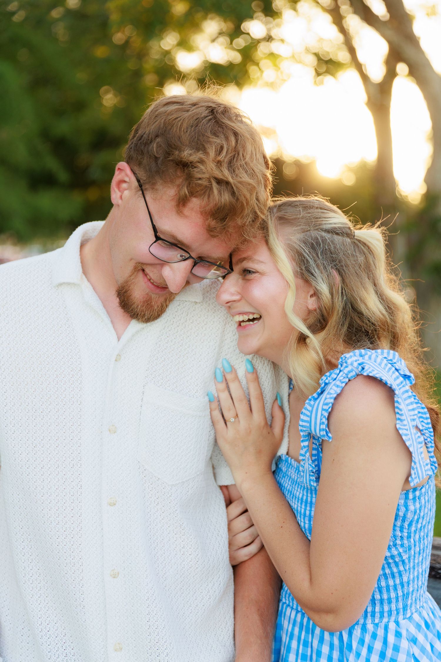 A smiling couple shares a joyful moment outdoors at golden hour, woman in blue dress rests hand on man's chest.