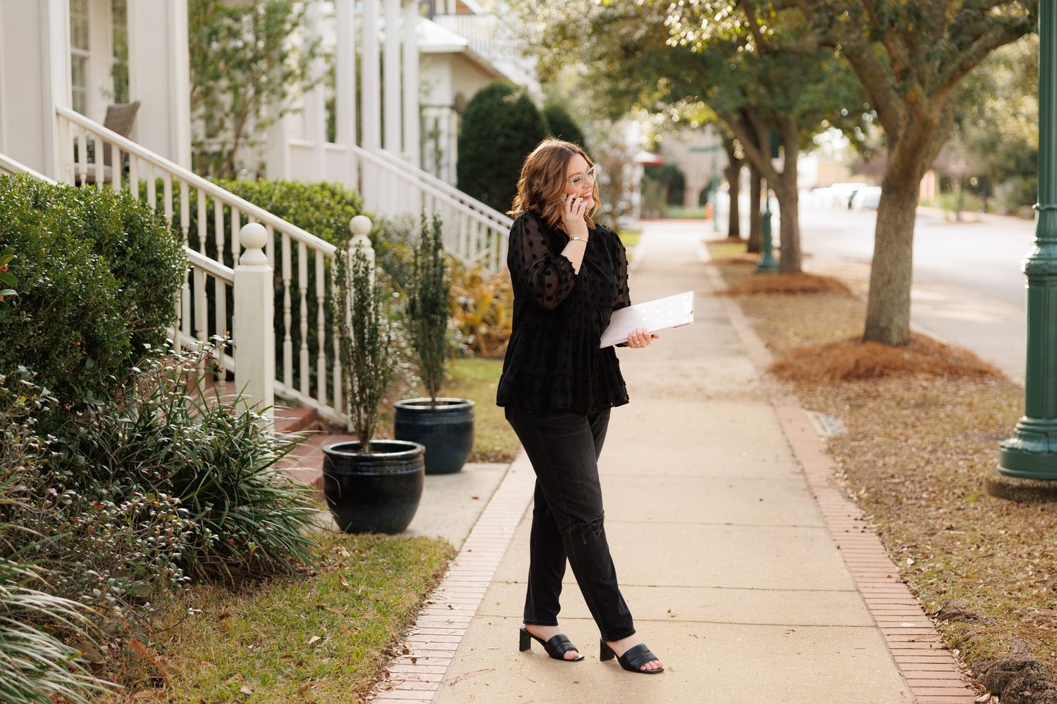 Woman in black suit walking on sidewalk talking on phone while holding documents in a suburban neighborhood.