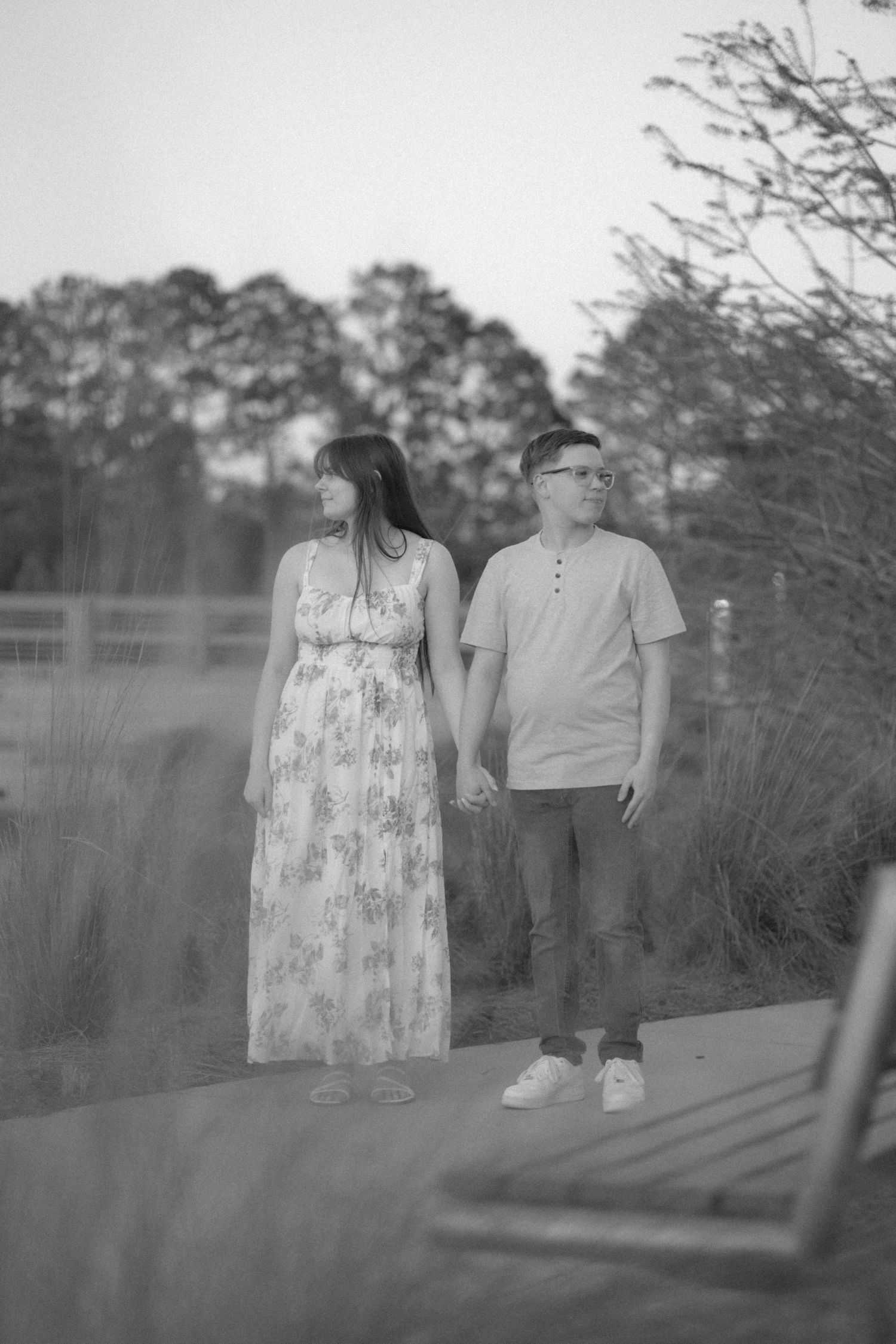 Black and white photo of a young couple holding hands on a path surrounded by tall ornamental grasses.