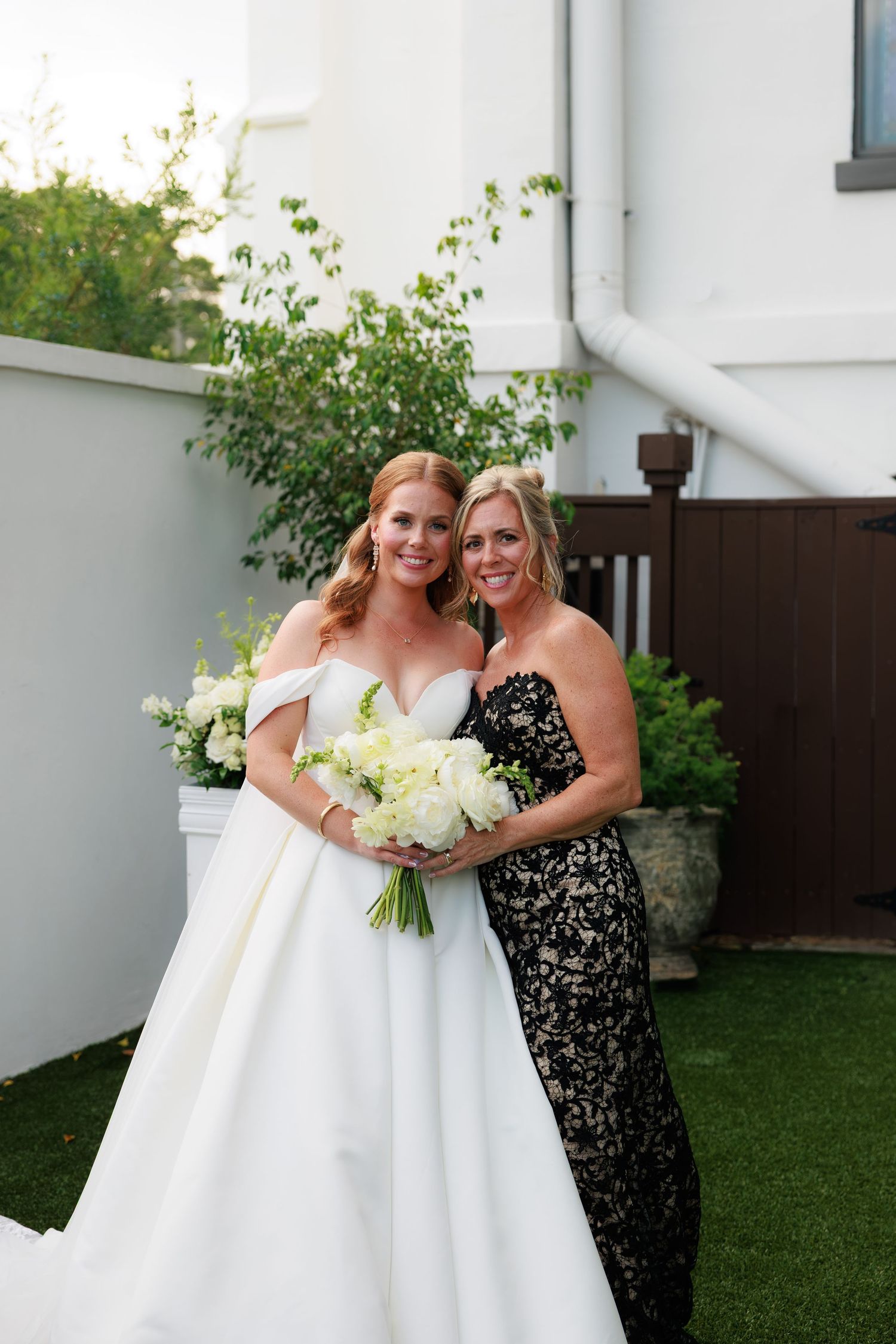 Bride in white off-shoulder gown holding white bouquet poses with woman in black lace dress outdoors.