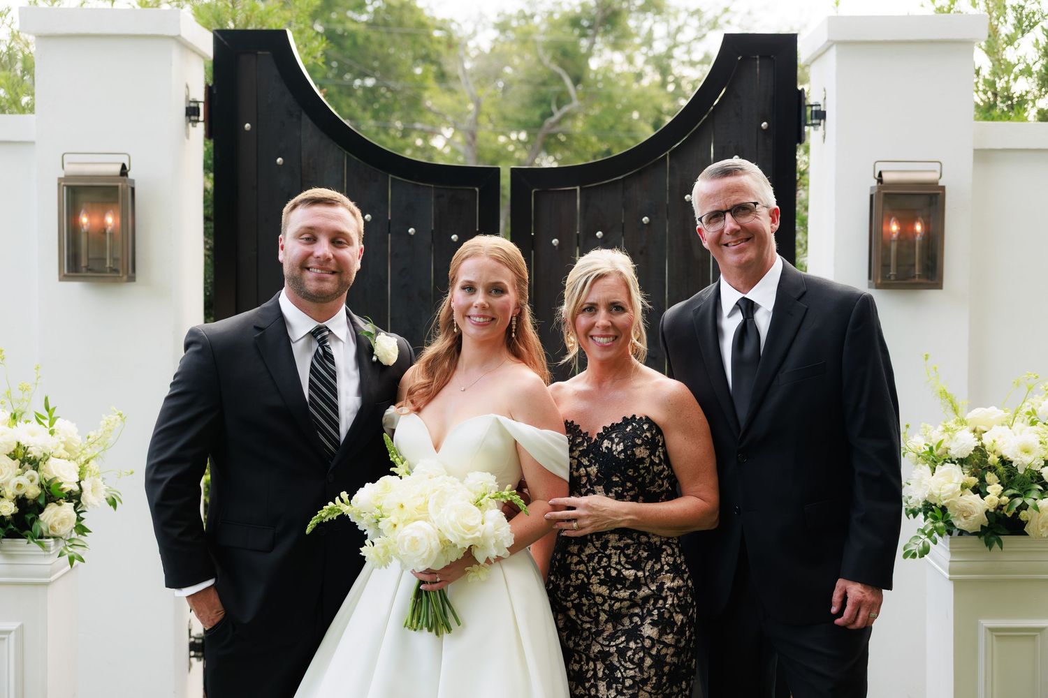 Bride in white gown holding bouquet poses with groom and family in front of elegant black gates at outdoor wedding.