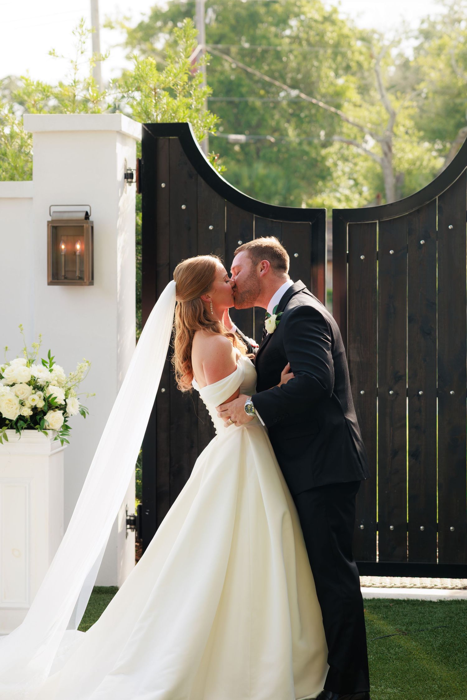 Bride and groom share first kiss at outdoor ceremony by elegant dark wooden gates with white floral arrangements.