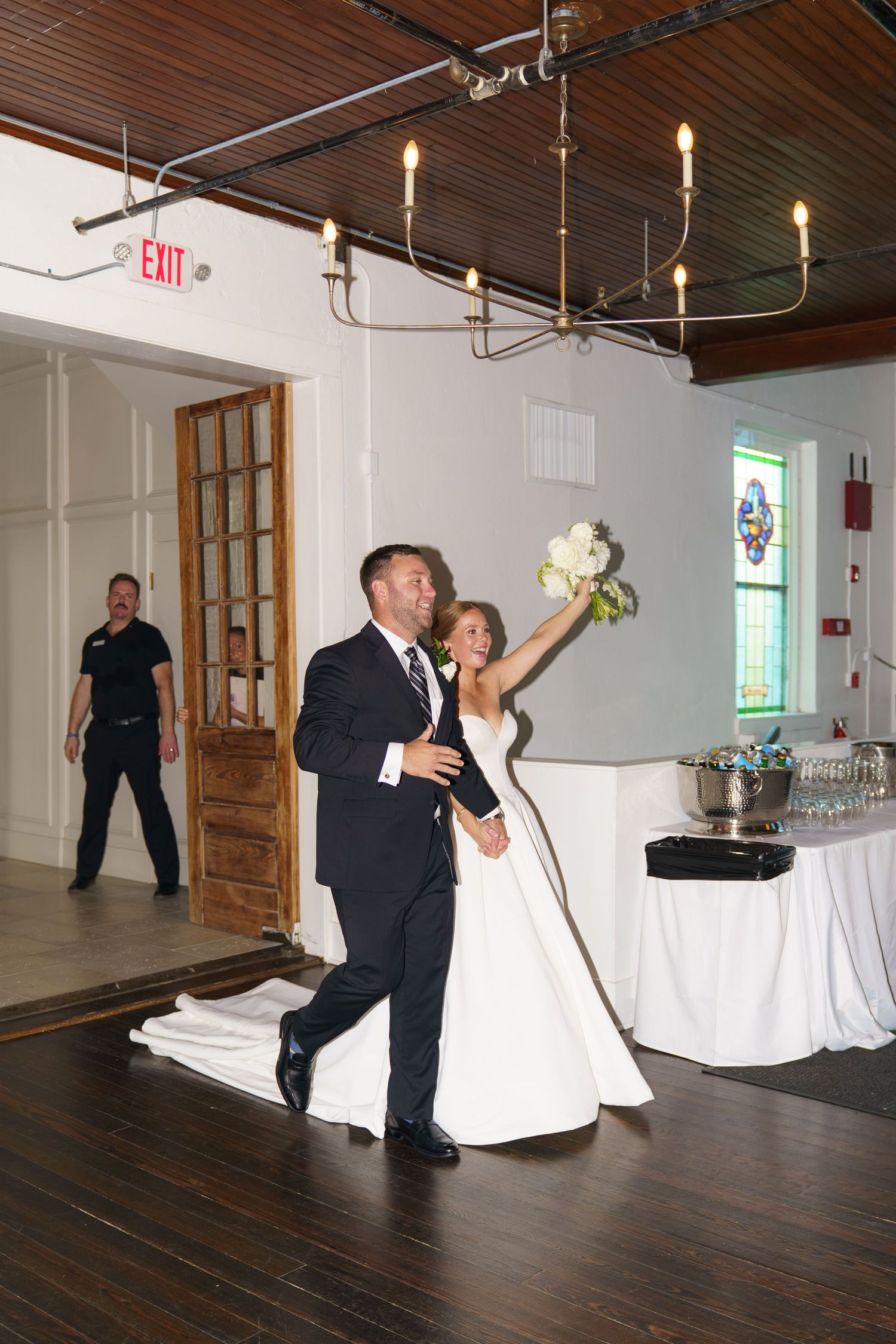 Bride raises bouquet triumphantly as groom escorts her into elegant wedding reception venue with stained glass windows.