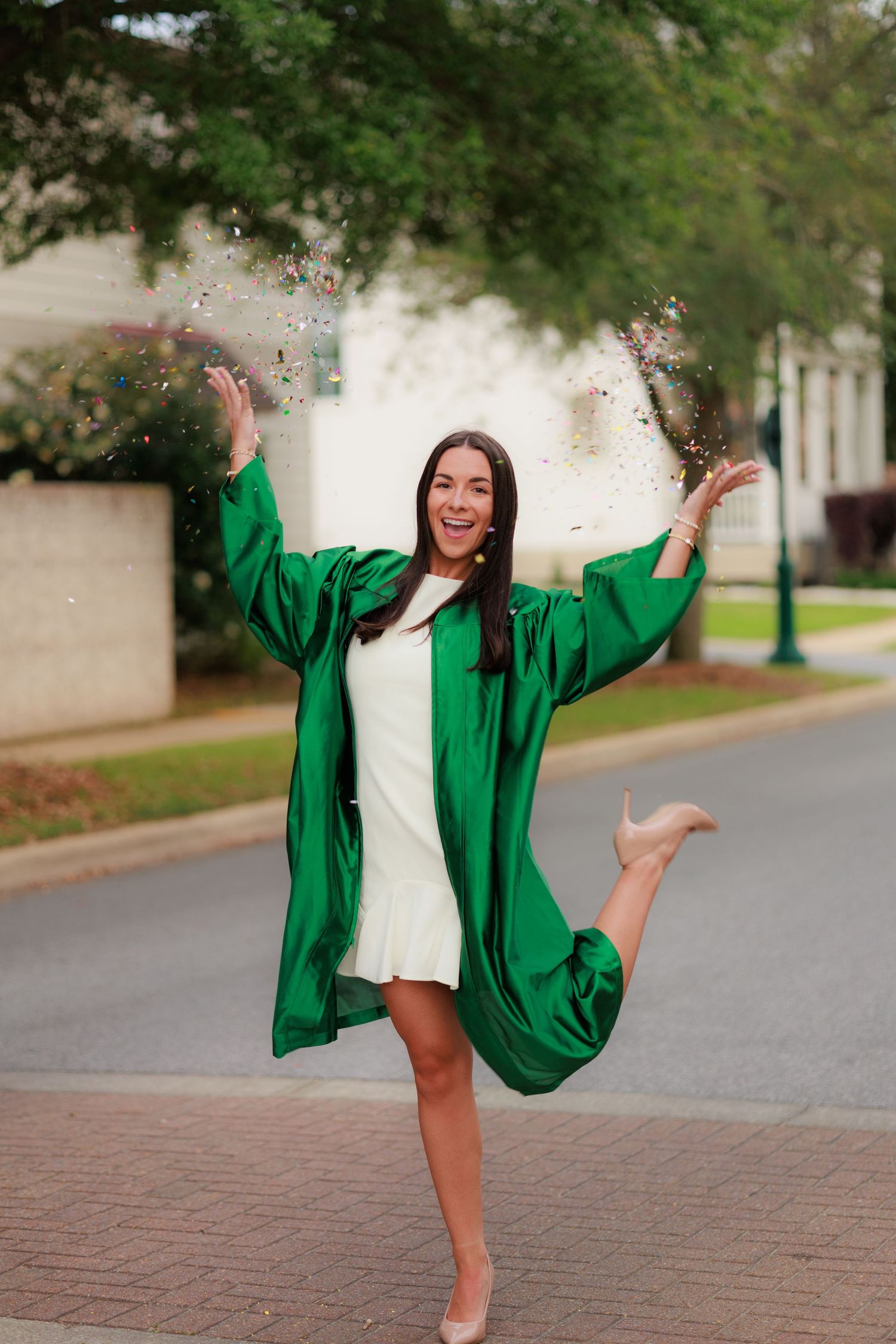 A joyful woman in a green graduation gown tosses confetti while balancing on one leg outdoors.