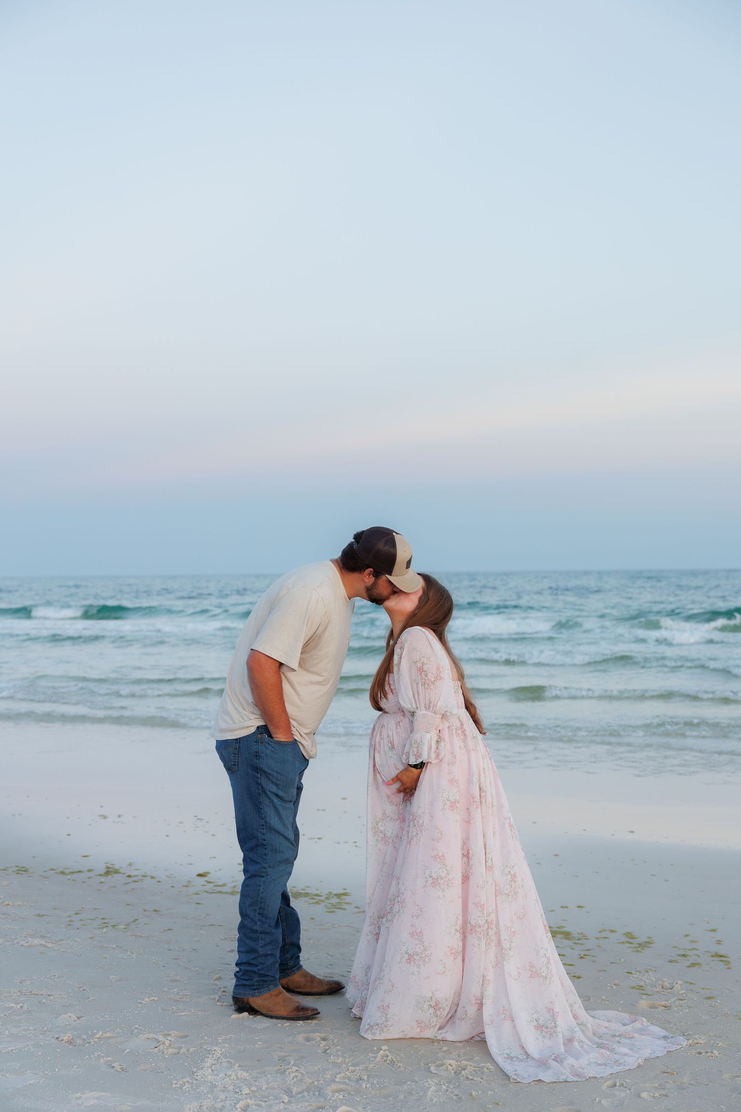 Pregnant couple sharing a romantic beach kiss at dusk, woman in flowing floral maternity gown by the ocean.