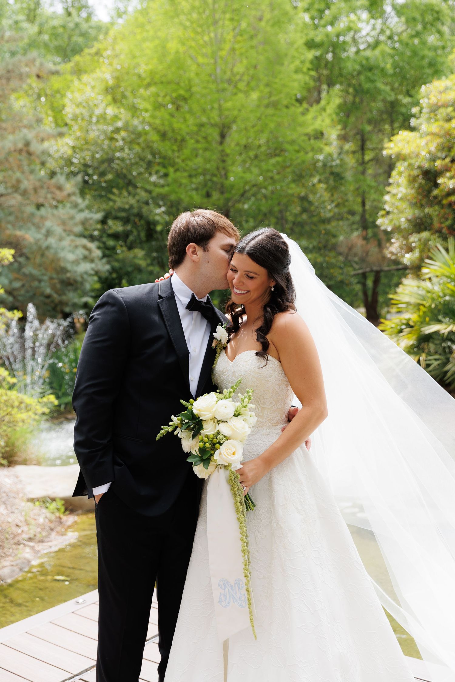 Groom whispering to laughing bride on wooden deck surrounded by lush green garden.